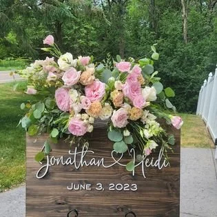 A wooden sign with white cursive text reading 'Jonathan & Heidi,' and the date June 3, 2023, adorned with a large floral arrangement of pink, white, and peach roses and greenery outdoors.