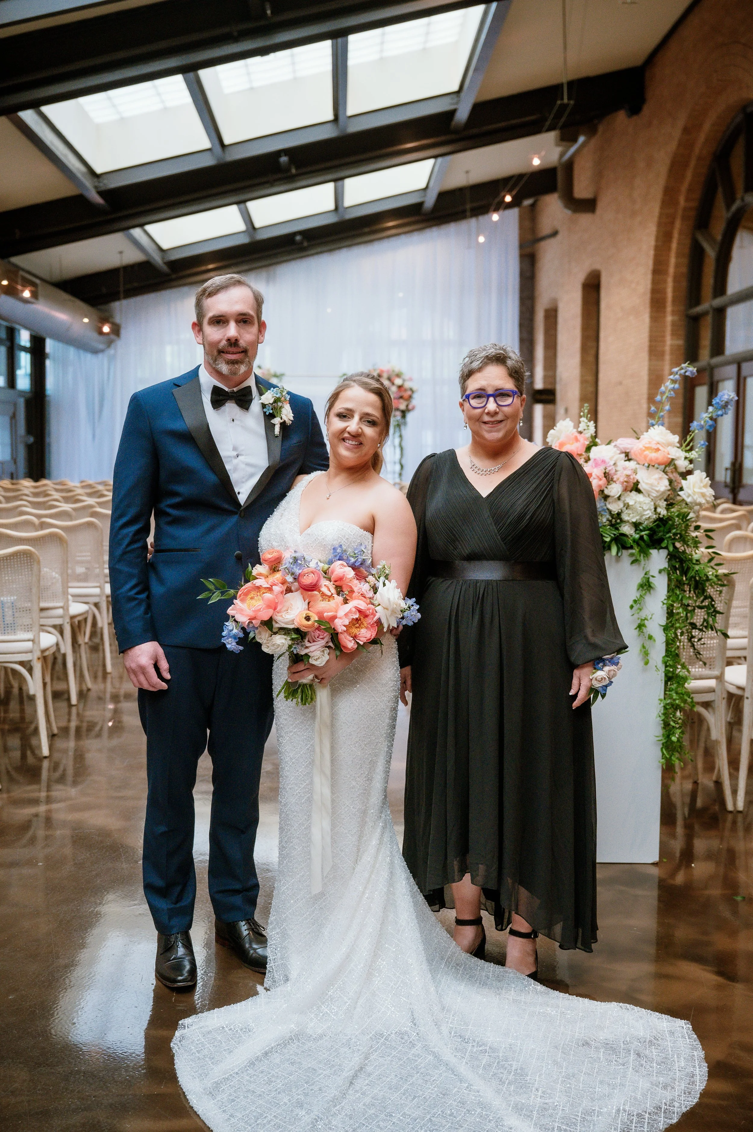 A wedding scene with three people posing indoors; a groom in a blue tuxedo and bow tie, a bride in a white wedding gown holding a bouquet of flowers, and a woman in a black dress with glasses. There are floral arrangements and chairs in the background.