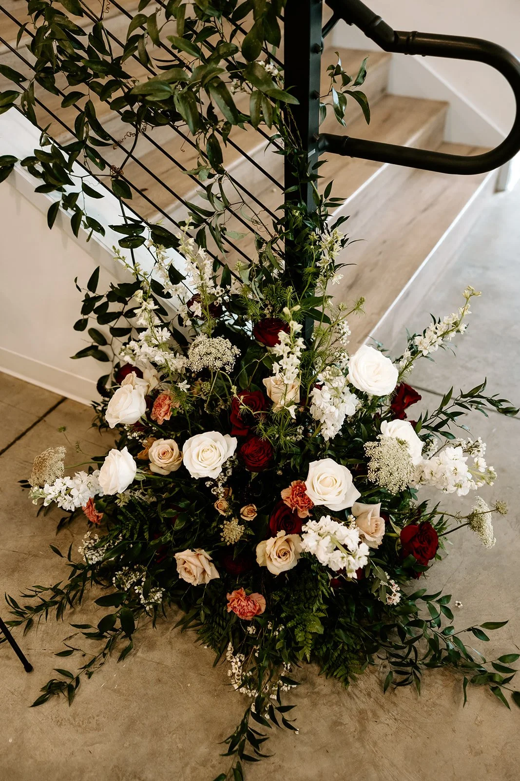 A floral arrangement with white, red, and pink roses, white stock, and greenery, placed on a concrete floor next to a black metal railing and a staircase.