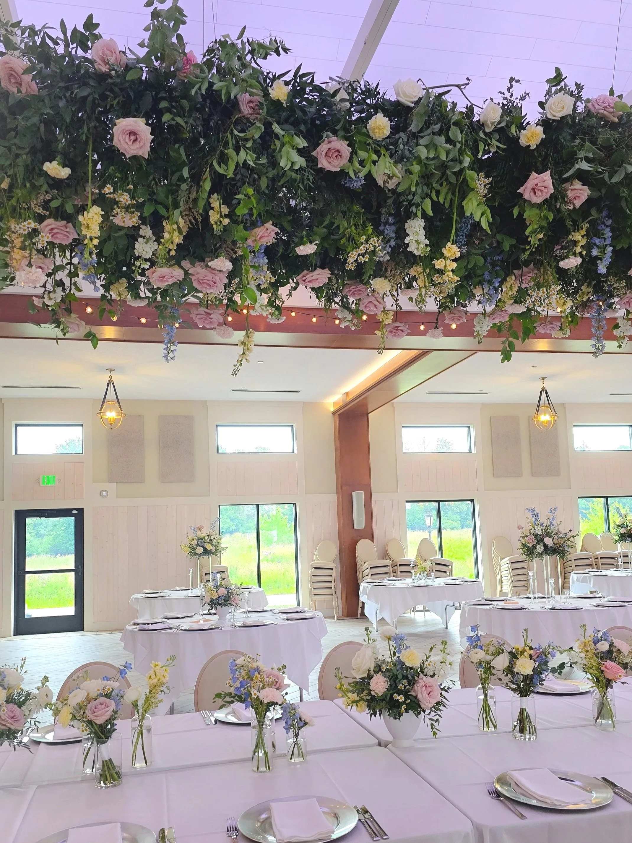 Indoor wedding reception with round tables covered in white tablecloths, decorated with floral centerpieces in vases, set with plates, napkins, and silverware. Chandeliers and string lights hang from the ceiling, and large windows provide natural lig