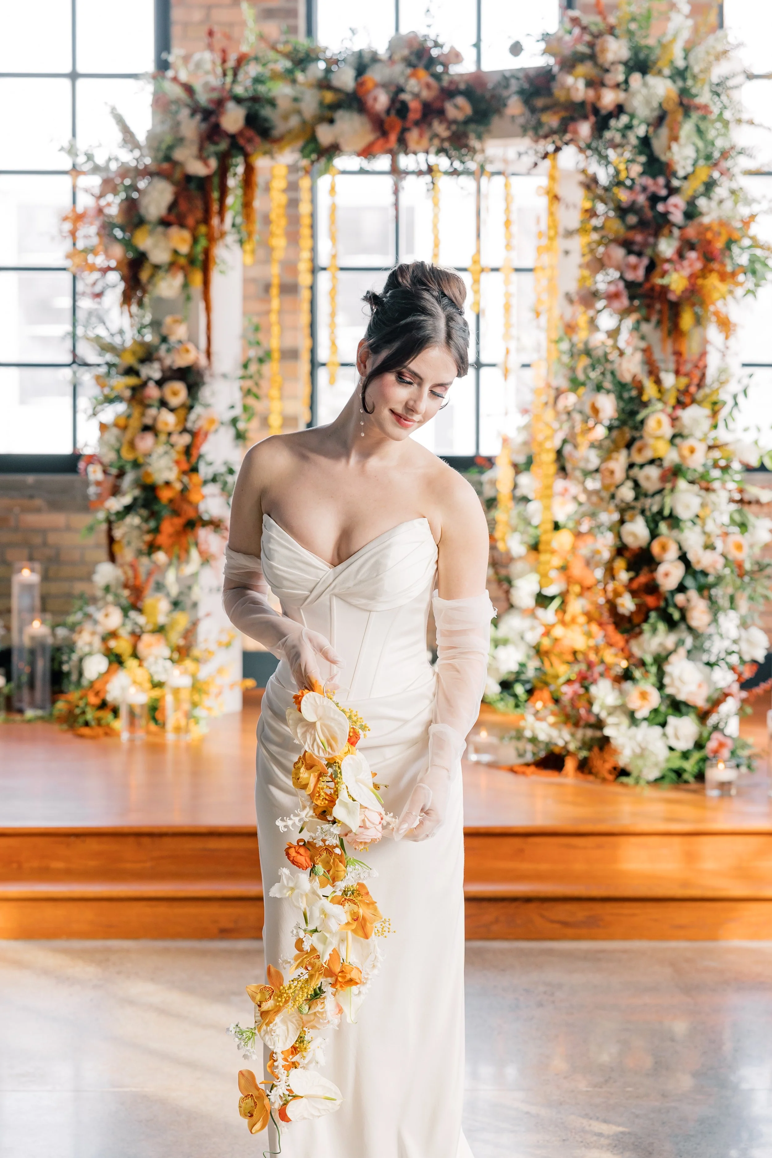 A woman in a white wedding dress holding a cascading floral bouquet stands in front of an arch made of flowers and greenery at a decorated wedding venue.