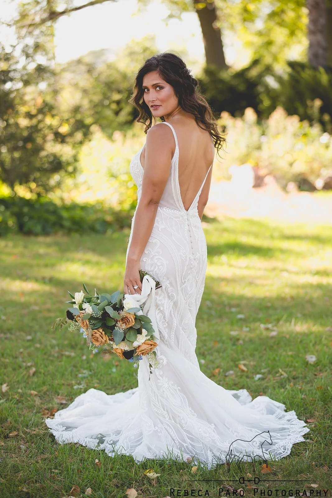 A woman in a white wedding gown holding a bouquet of flowers outdoors, with trees and greenery in the background.