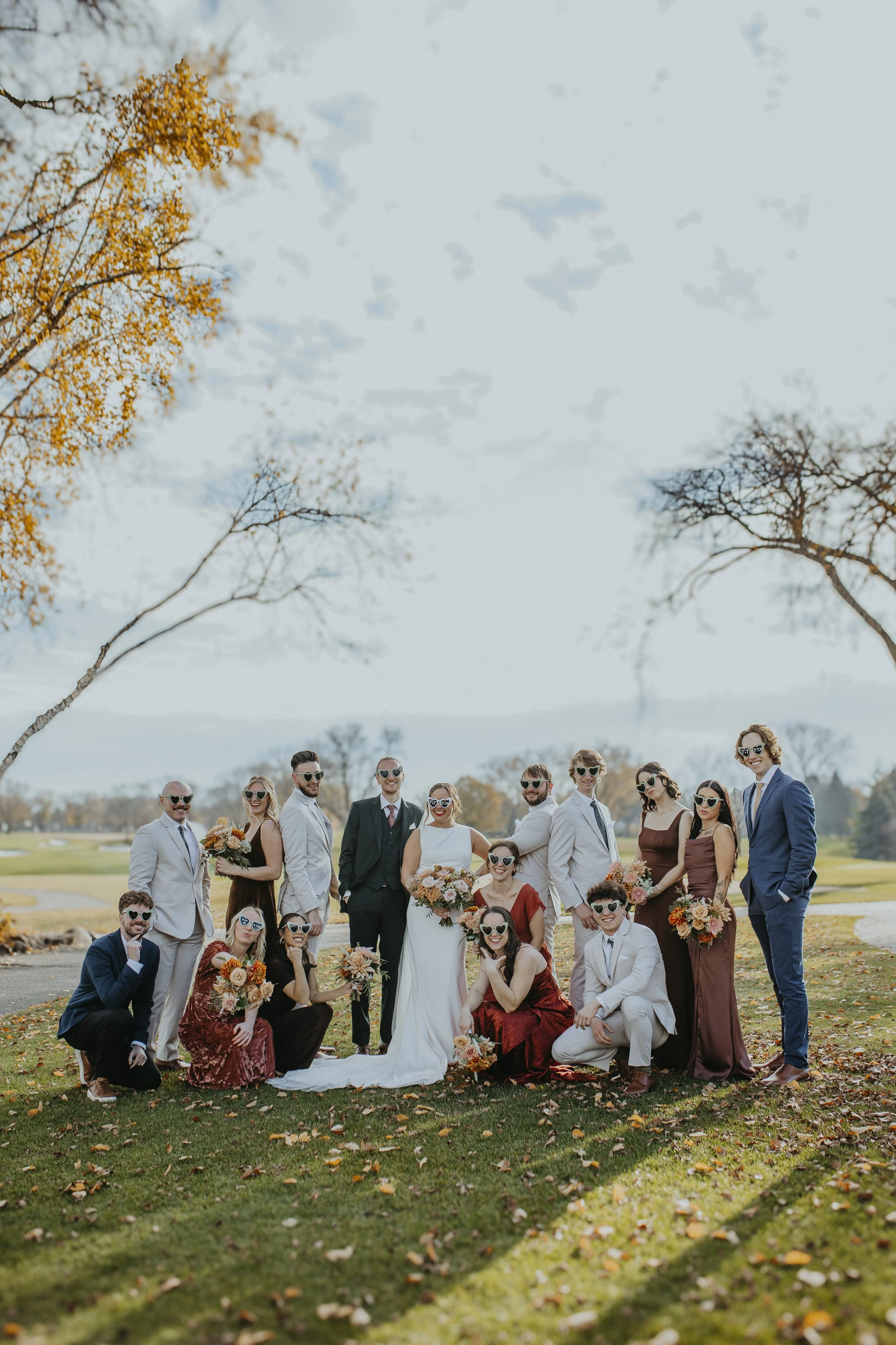 A group of people dressed in formal wedding attire, wearing heart-shaped sunglasses, posing outdoors in a park with trees and fallen leaves, under a partly cloudy sky. The group includes men, women, and children holding bouquets and smiling.