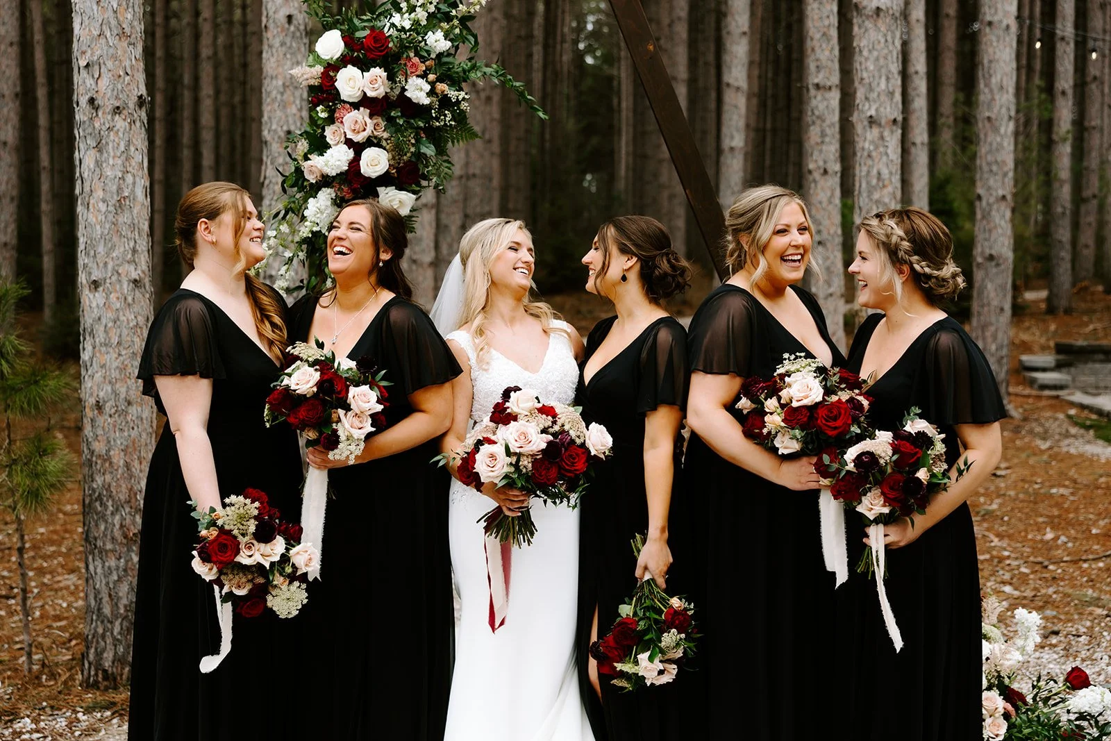 A group of six women, including a bride in a white wedding dress and five bridesmaids in black dresses, standing outdoors in a forested area with tall trees. They are smiling and holding bouquets of red, white, and blush roses with greenery.