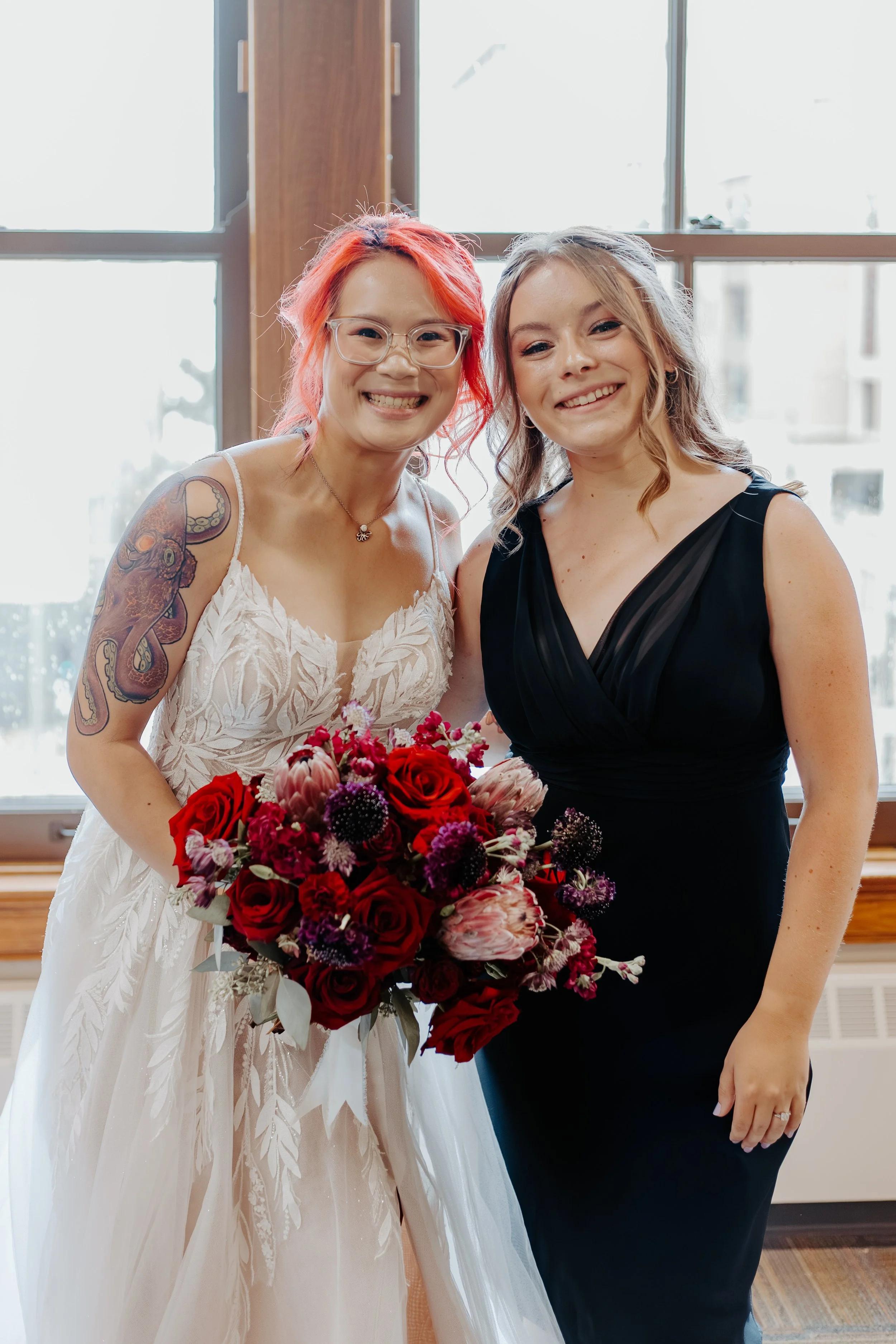 Two women smiling inside a room with large windows. One woman is wearing a white wedding dress and glasses, holding a bouquet of red, pink, and purple flowers. The other woman is in a black dress with wavy blonde hair.