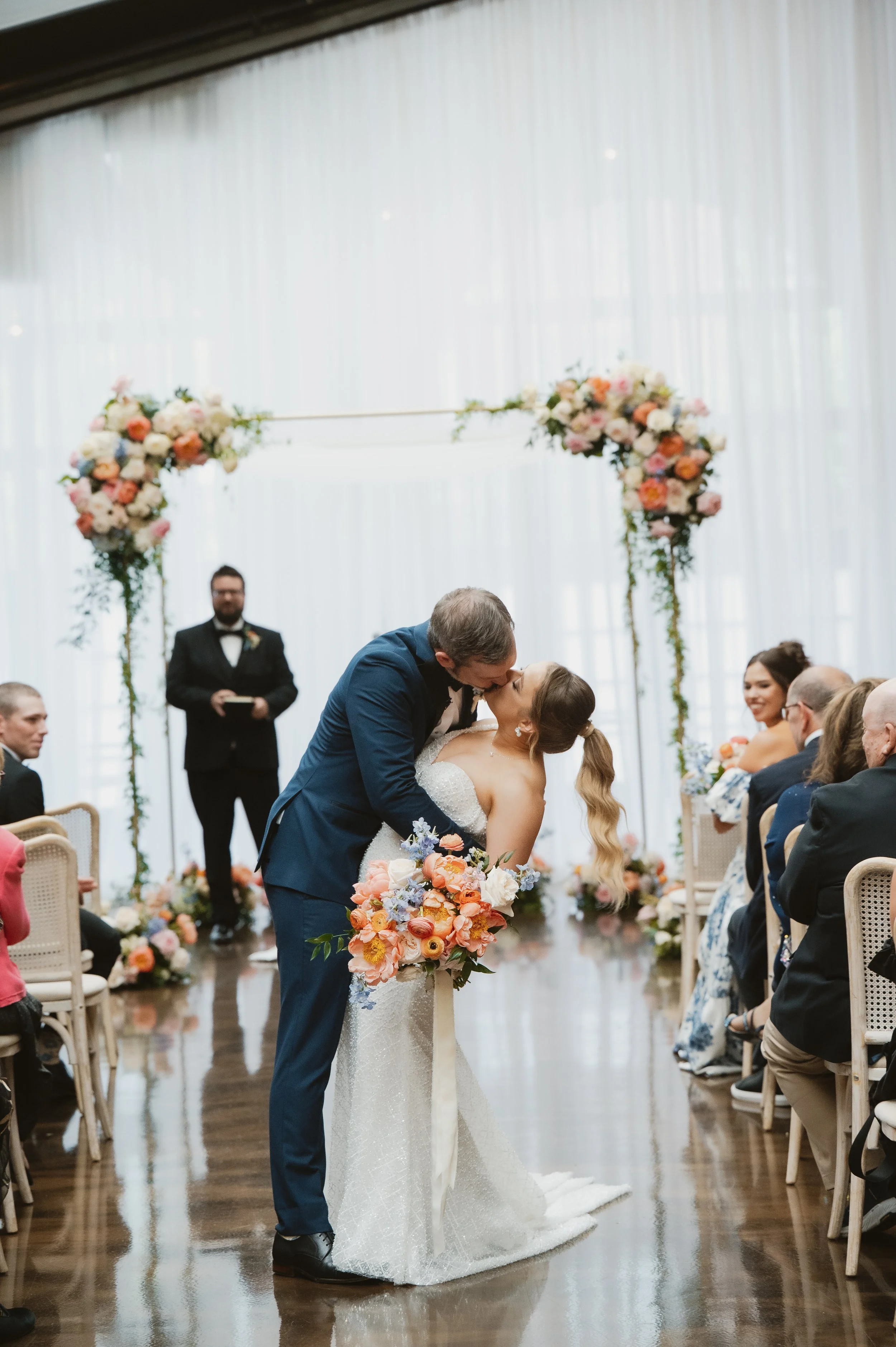 A bride and groom kiss during their wedding ceremony in a decorated indoor venue with floral arrangements and seated guests.