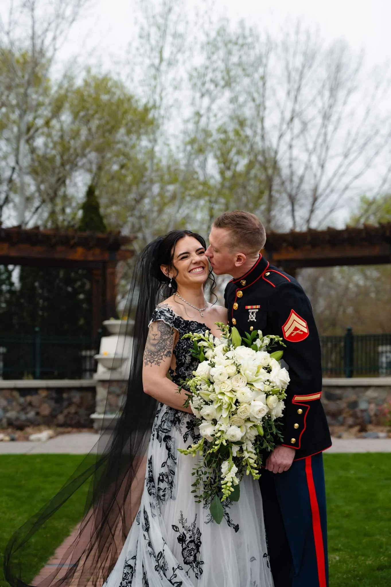 A woman in a wedding dress holding a bouquet of white flowers with a man in a U.S. Marine dress uniform kissing her on the cheek outdoors, with trees and a wooden structure in the background.