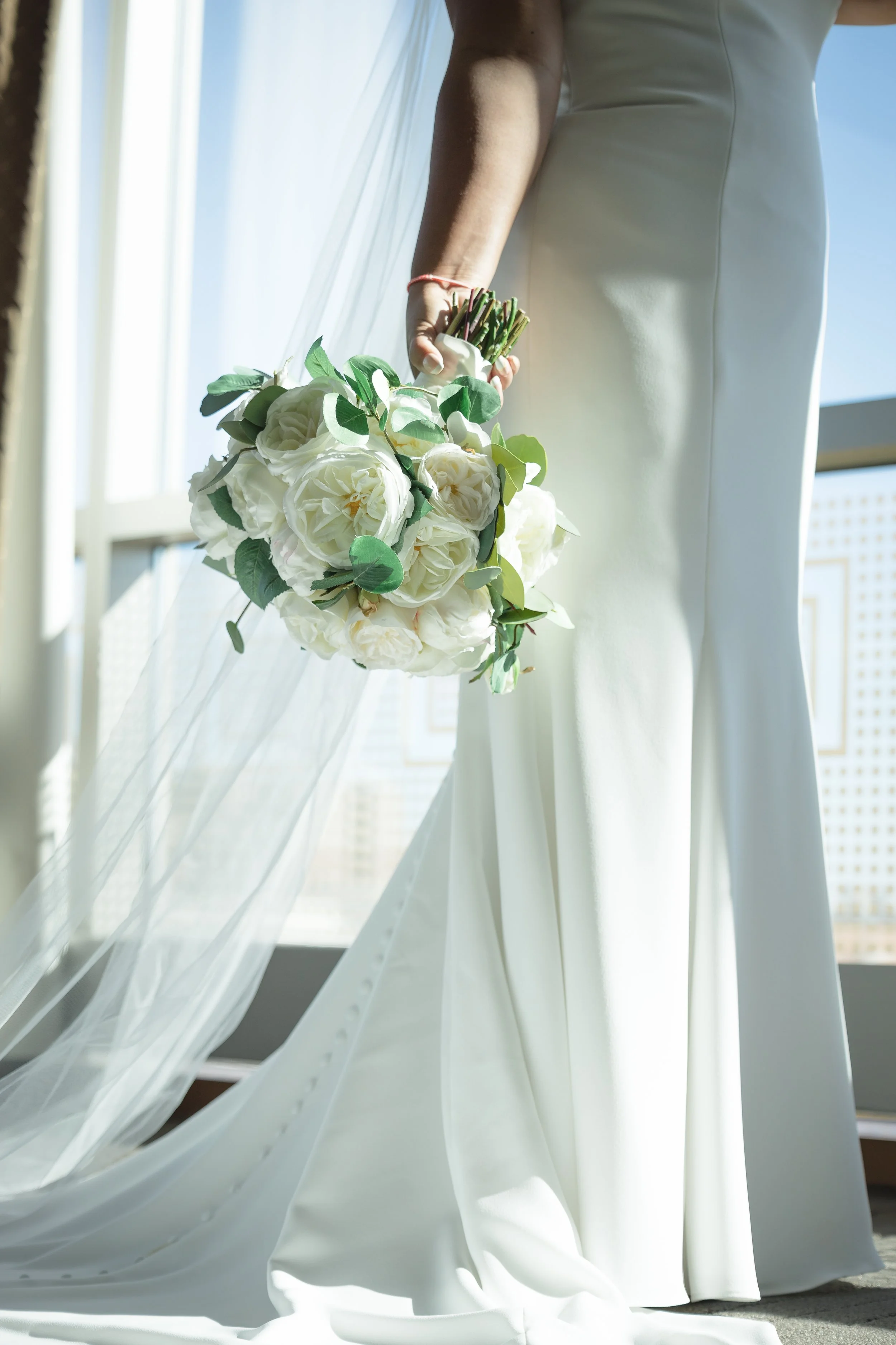 A bride holding a bouquet of white roses and green leaves, standing in front of large window with sheer curtains, wearing a white wedding gown.