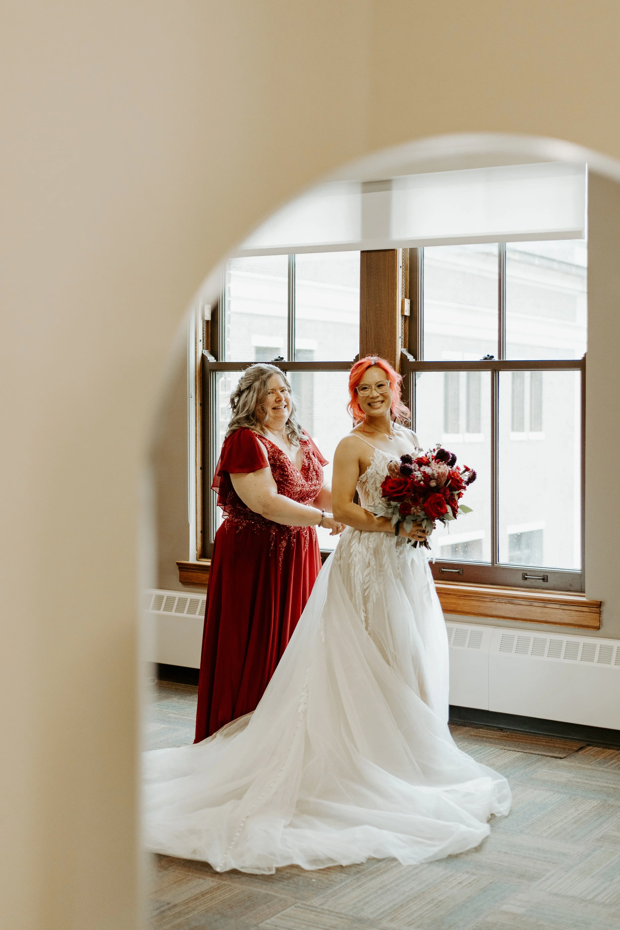 A bride in a white wedding gown holding a bouquet of red, pink, and purple flowers, smiling, with an older woman in a red dress standing next to her, inside a room with large windows and wooden window frames.