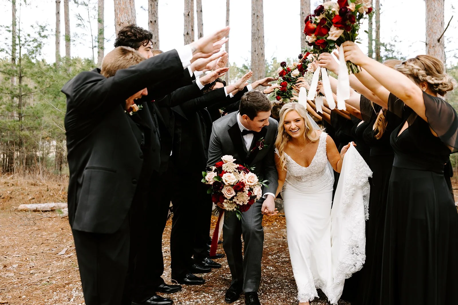 Bride and groom walking through a tunnel formed by wedding guests holding bouquets and hands in an outdoor forest setting, during a wedding ceremony.