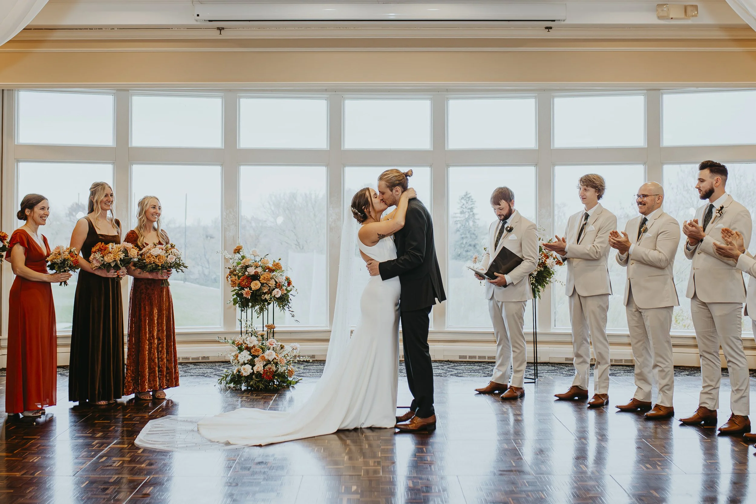 A wedding ceremony with a bride and groom kissing in front of a group of onlookers. The bride is dressed in a white gown, and the groom is in a black suit. Bridesmaids and groomsmen are on either side, with the bridesmaids holding bouquets of flowers. The setting is indoors with large windows and natural light.
