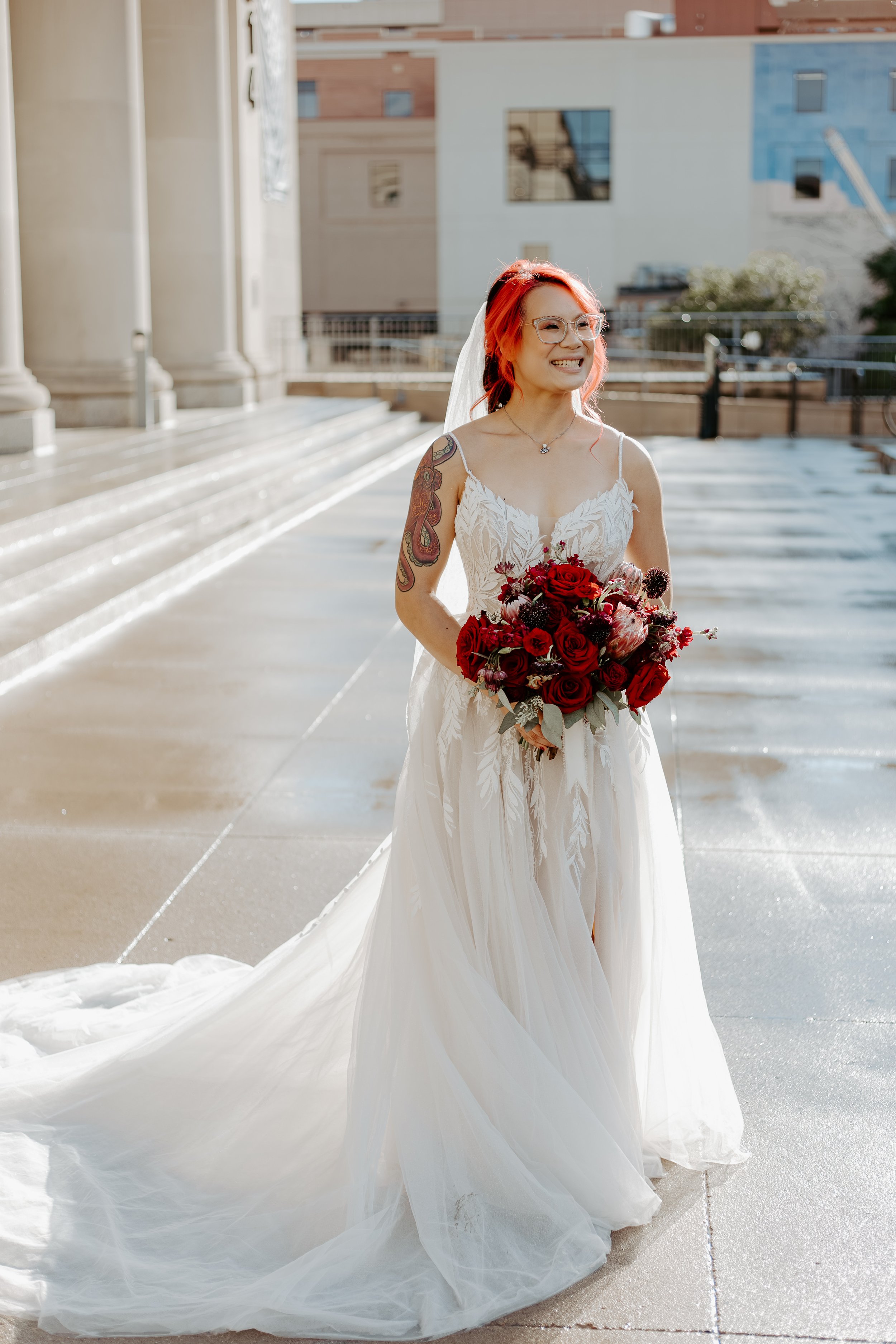 A person wearing a white wedding dress with spaghetti straps, holding a bouquet of red and dark purple flowers, standing outdoors on a sunny day. They have red hair, glasses, and tattoos on their left arm, and are smiling.