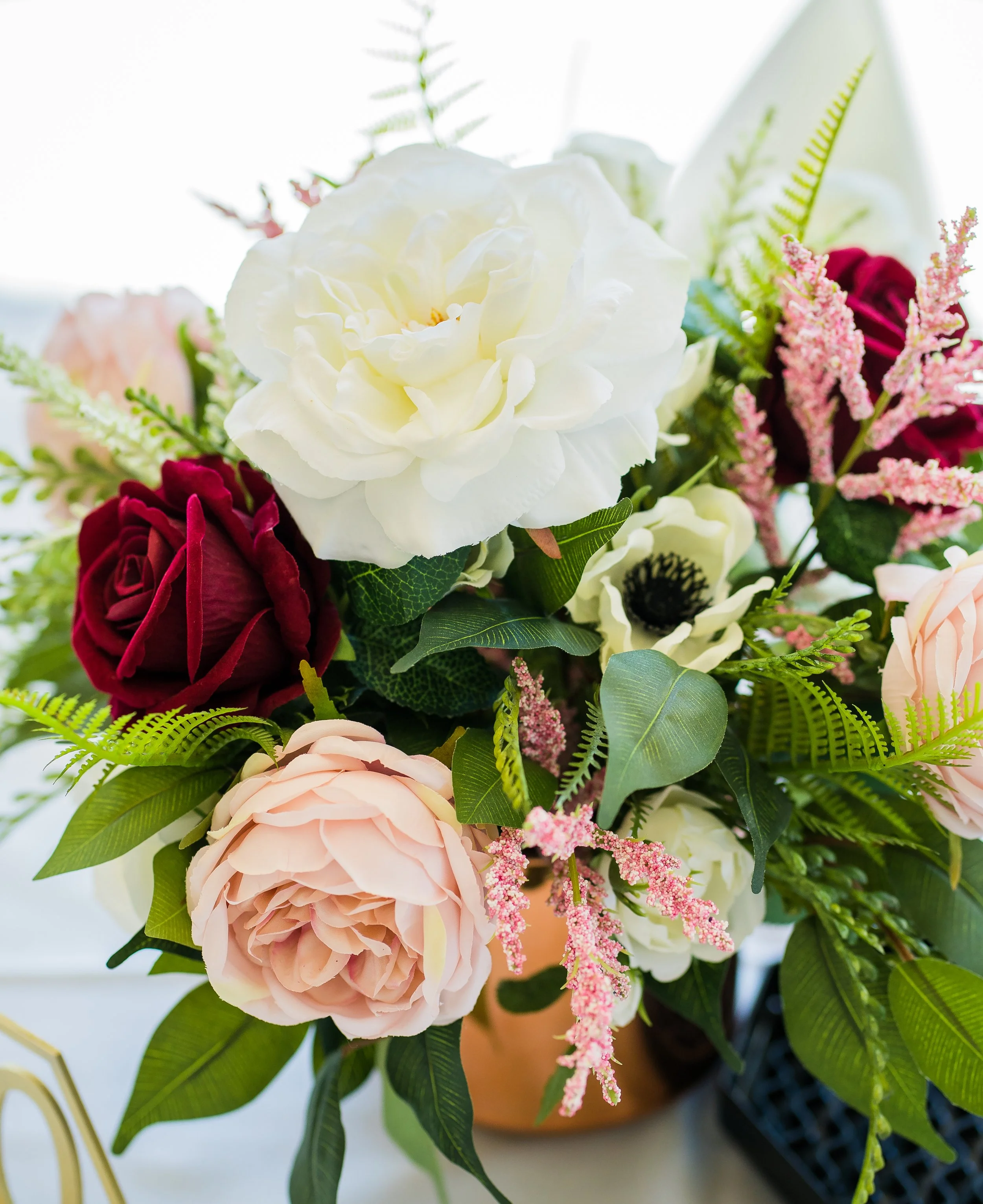 Close-up of a colorful flower arrangement with white, pink, and red flowers and green foliage in a tan vase placed on a white surface.