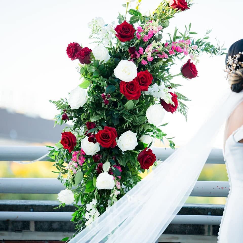 A large wedding floral arrangement with red, white, and pink roses and greenery, next to a woman's shoulder with a white dress and veil.