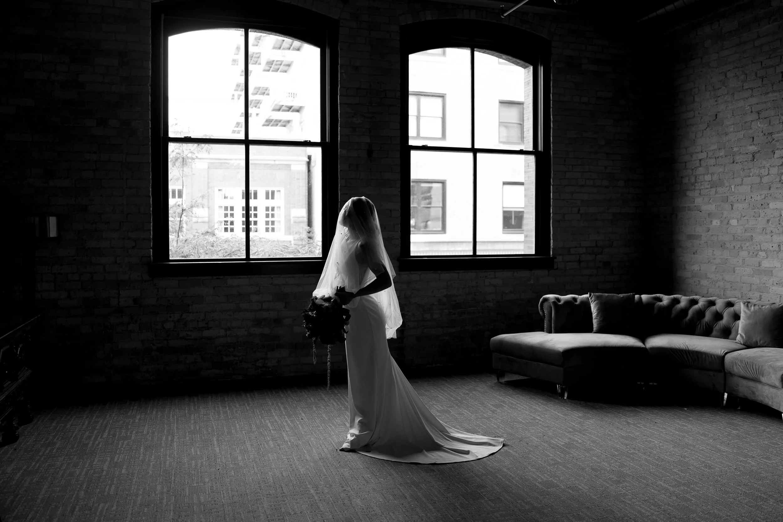 A black-and-white photo of a woman in a long dress and veil holding a bouquet, standing in a room with large windows and brick walls.