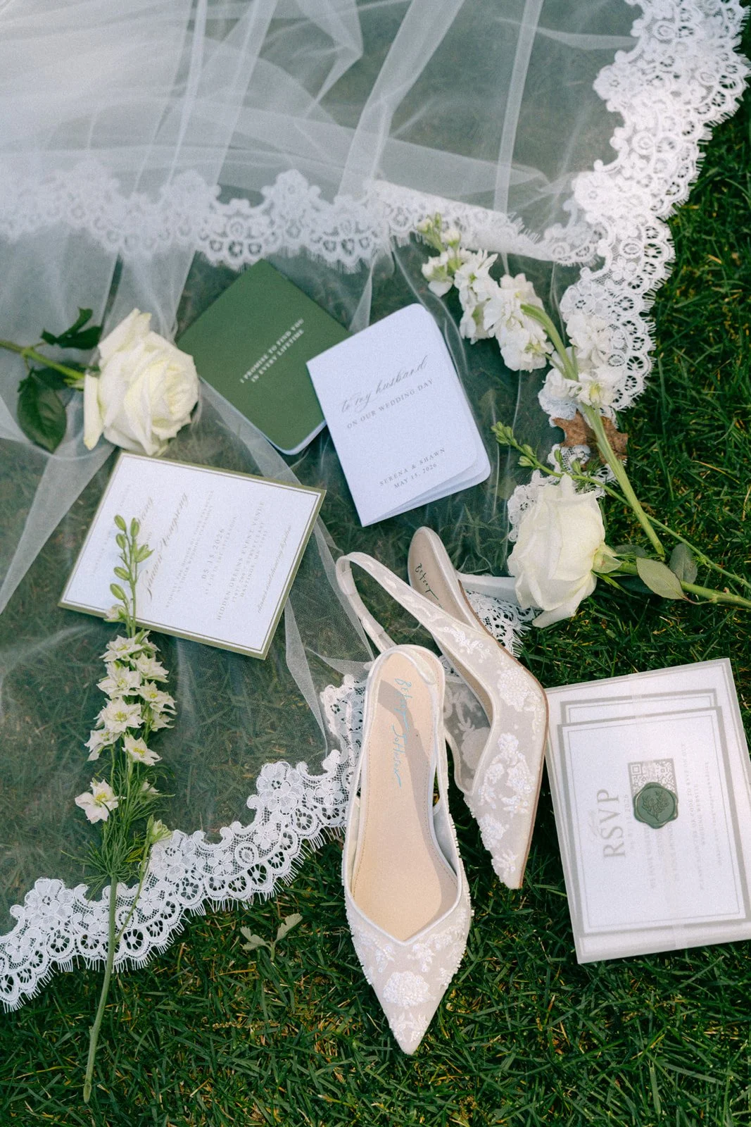 Wedding shoes, flowers, and RSVP cards on grass with lace fabric and greenery.