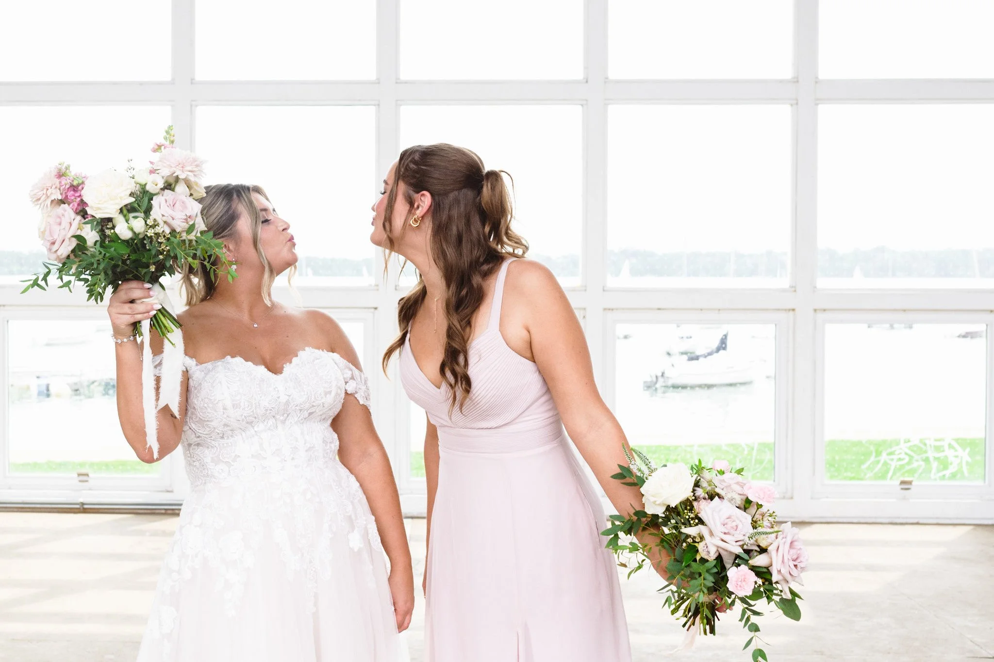 A bride and bridesmaid are about to kiss at a wedding, with large windows and a view of water and boats in the background.