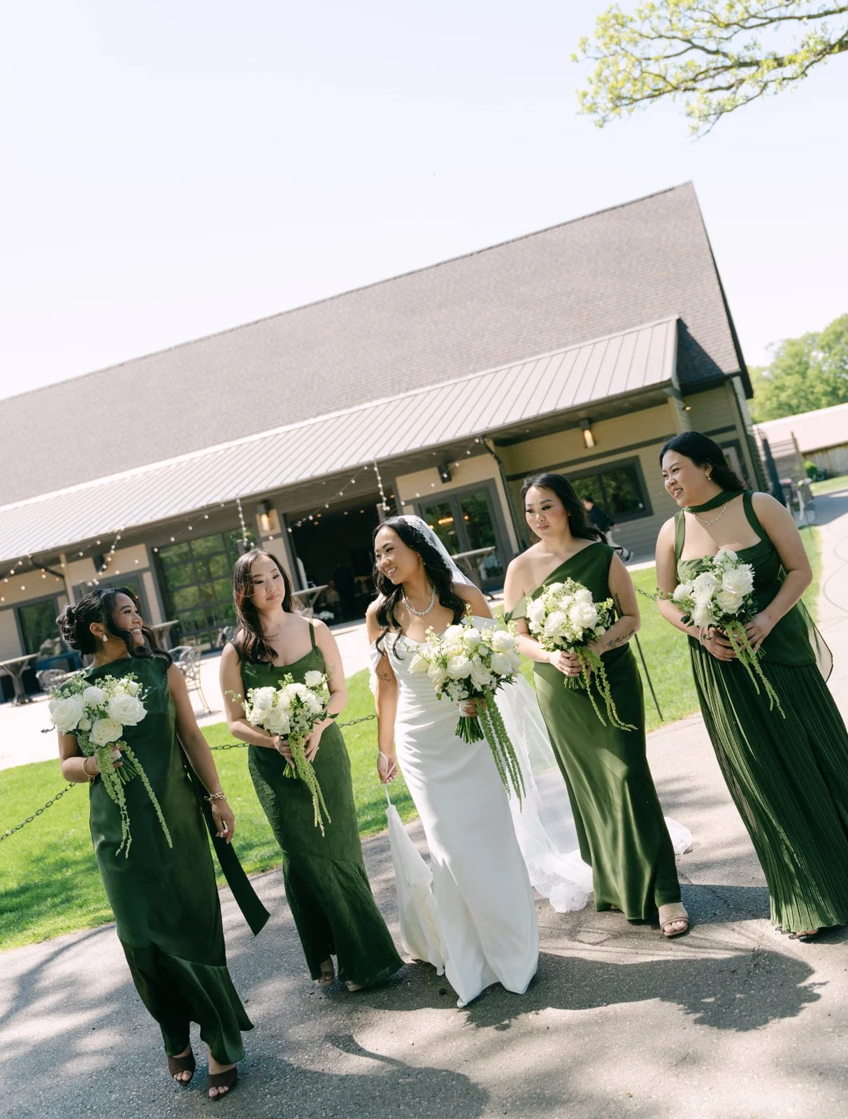 A bride and her bridesmaids walking outside of a building on a sunny day, all holding bouquets of white flowers, dressed in elegant green dresses except for the bride who is in a white gown.