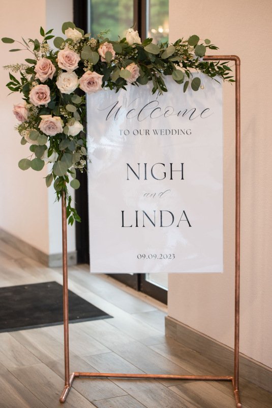 Wedding welcome sign on a copper stand decorated with pink and white roses and greenery, reading "Welcome to our wedding, Nigh and Linda, 09.09.2023."