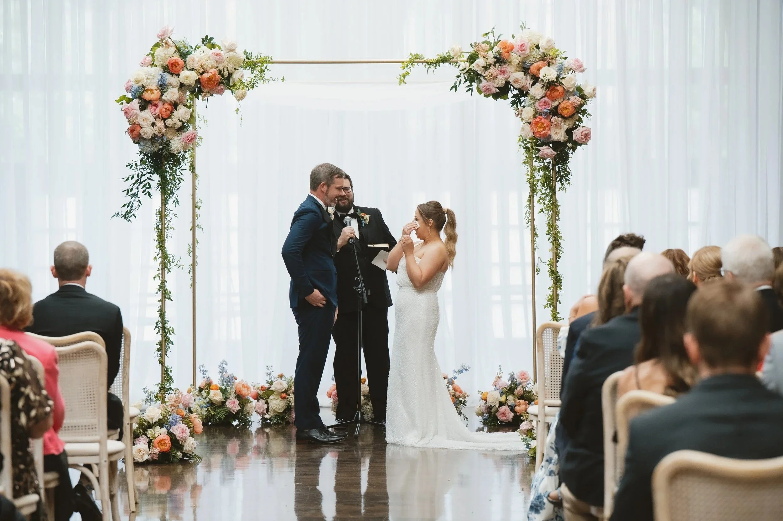 A wedding ceremony with a couple standing under a floral arch, exchanging vows, surrounded by seated guests in a bright indoor setting.