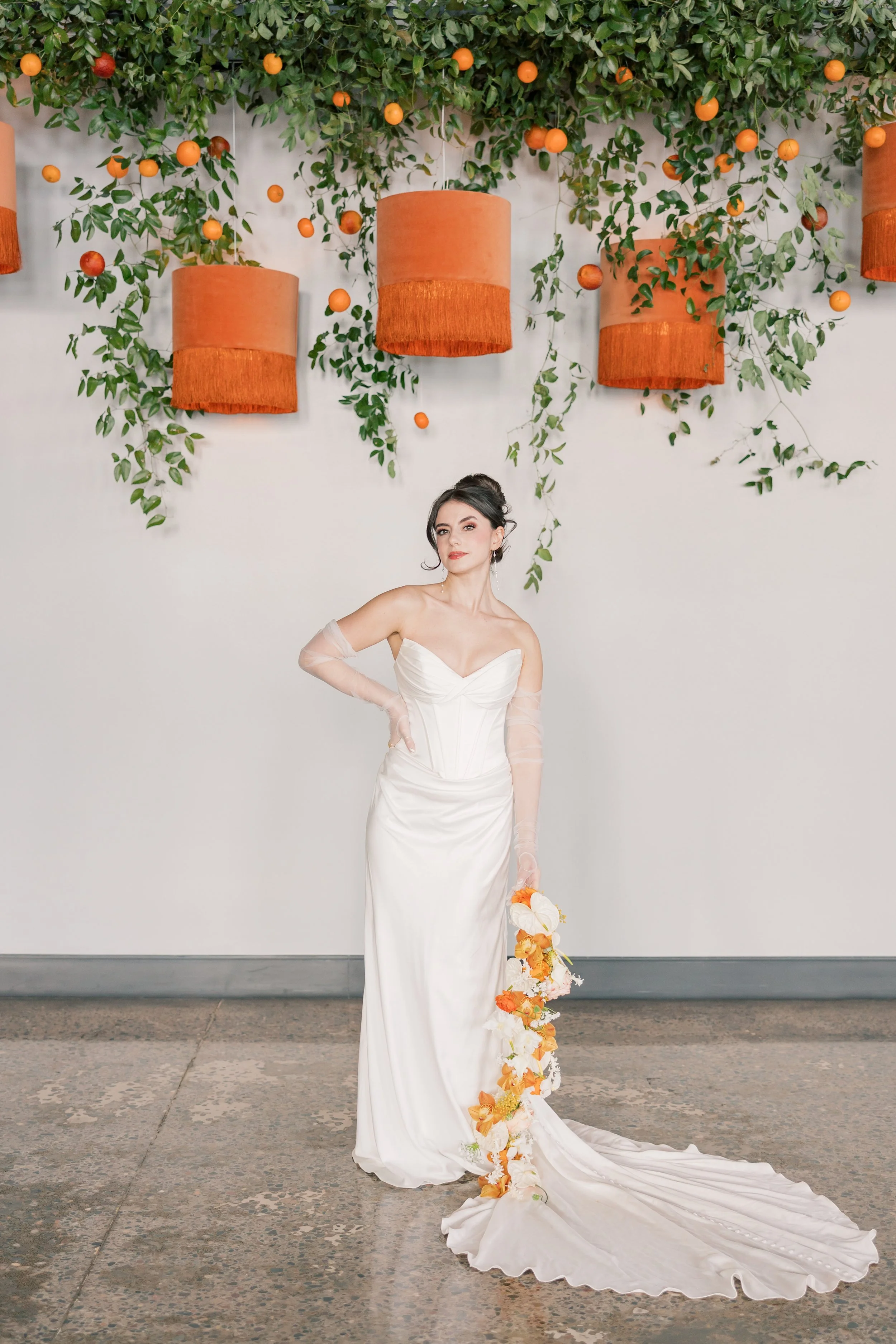 A woman in a long white wedding gown with sheer long sleeves, holding a bouquet of orange and white flowers, standing in front of a decorative green foliage wall with hanging orange lanterns.