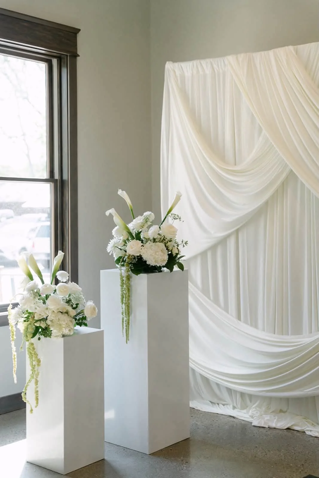 White floral arrangements on white pedestals in front of a beige wedding backdrop with draped fabric, near a window with gray trim.