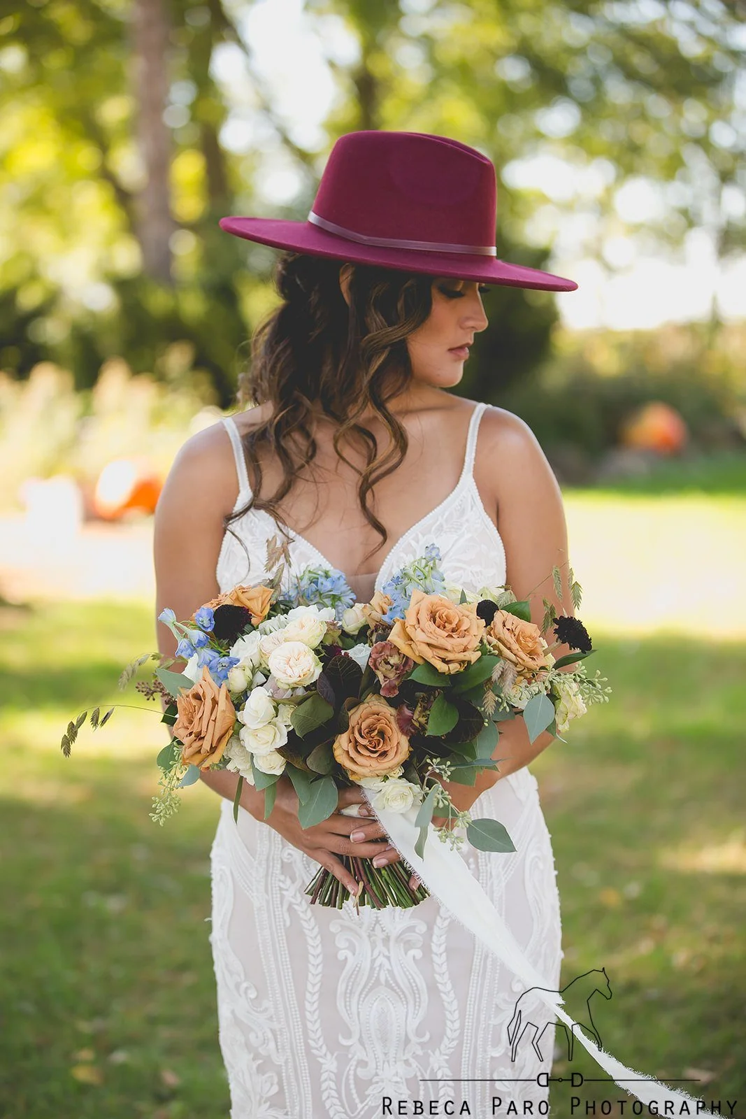 A woman with dark, curly hair wearing a maroon wide-brimmed hat and a white lace dress, holding a large bouquet of mixed flowers outdoors, with trees and greenery in the background.