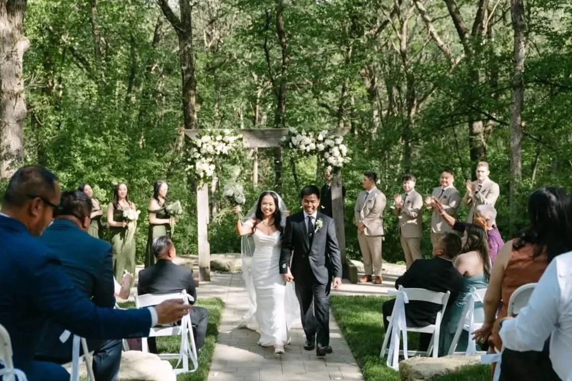 A wedding ceremony taking place outdoors in a wooded area with a couple walking down the aisle, surrounded by guests and wedding party, with a floral arch in the background.