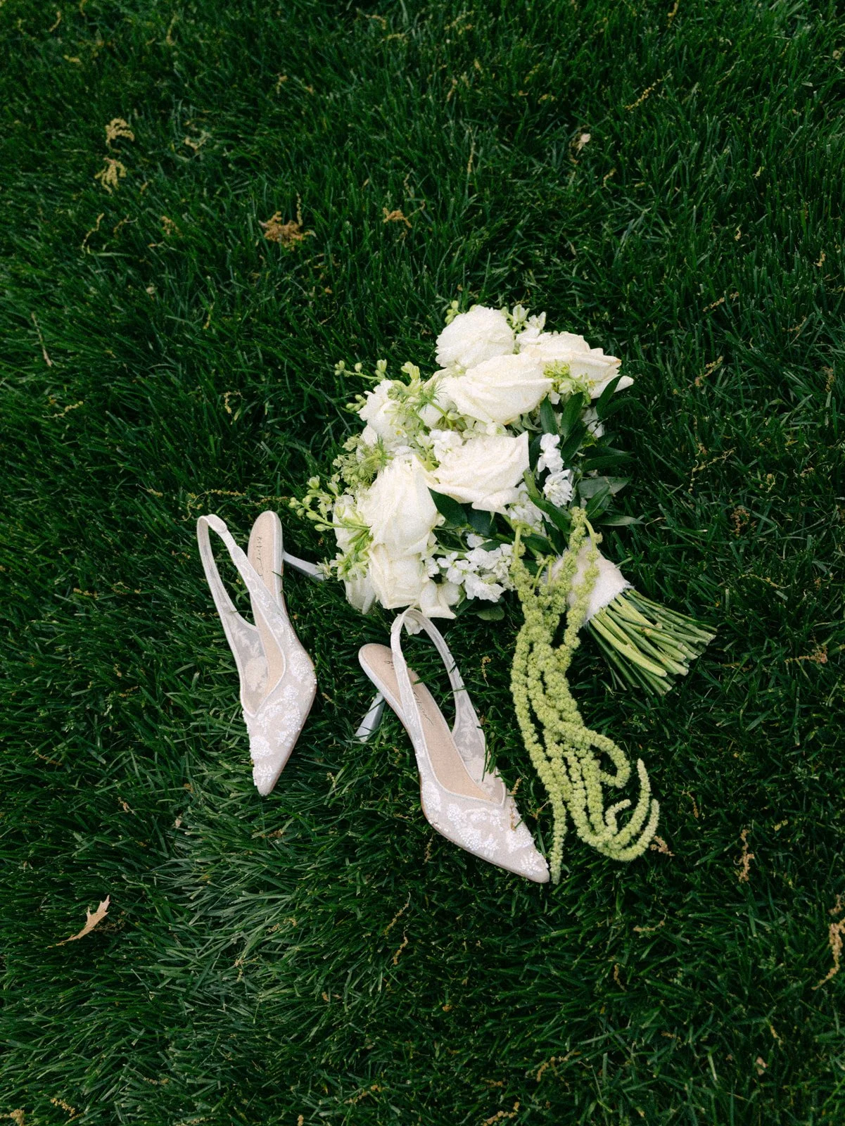 A bouquet of white flowers and wedding shoes on green grass.