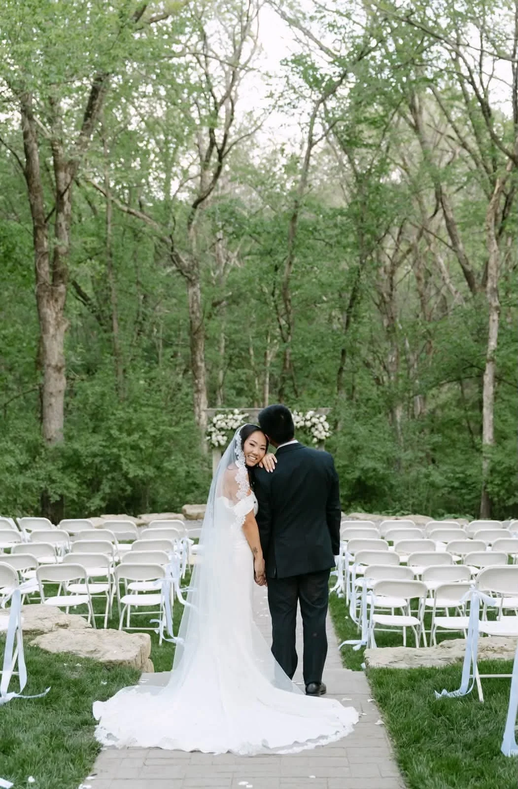 A bride and groom walking down the aisle in an outdoor wedding ceremony surrounded by trees, with chairs set up on both sides and floral decorations in the background.