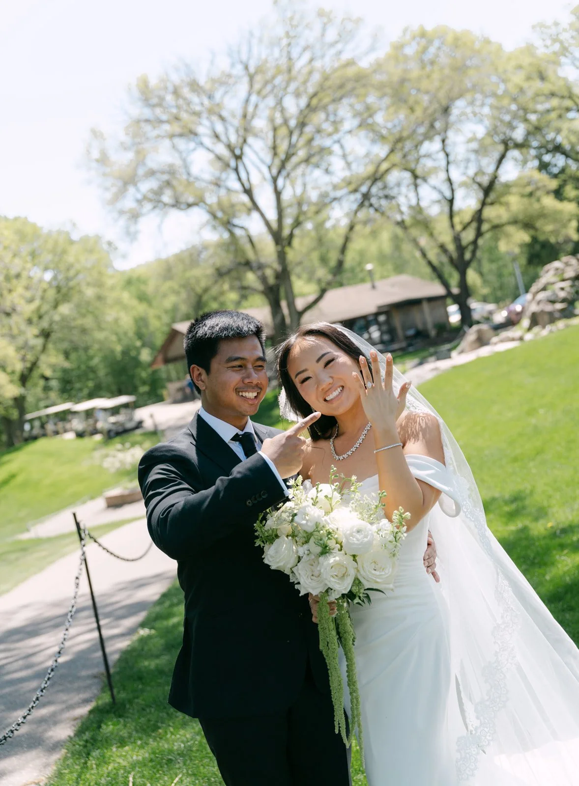 A newlywed couple in wedding attire, smiling and showing off their rings outdoors in a park-like setting with green grass, trees, and a building in the background.