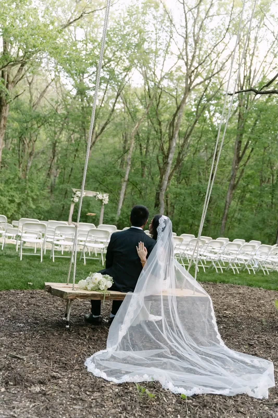 A bride and groom sitting on a wooden swing outdoors, surrounded by trees, with wedding chairs and an arch visible in the background, during a wedding ceremony.