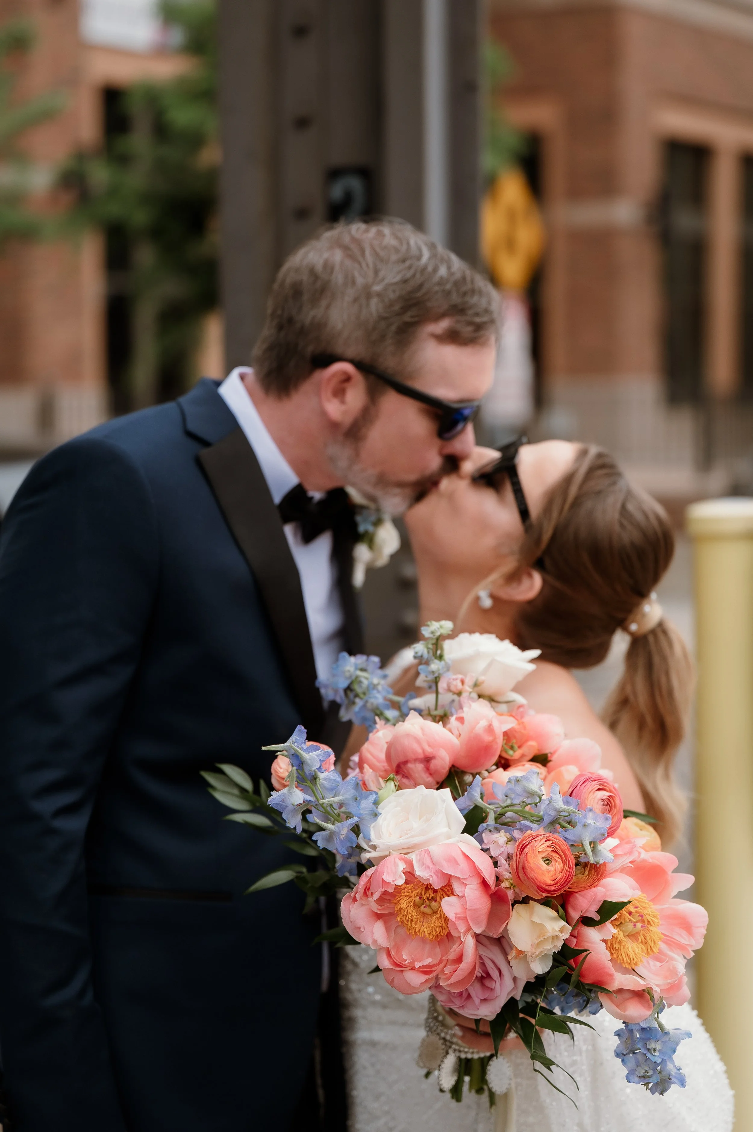 A couple sharing a kiss during a wedding, with the woman holding a large bouquet of pink, white, and blue flowers.