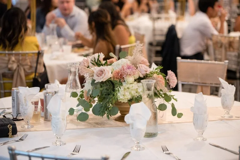 A decorated banquet table at a formal event with a large floral centerpiece in a gold container, surrounded by glassware, silverware, and napkins.