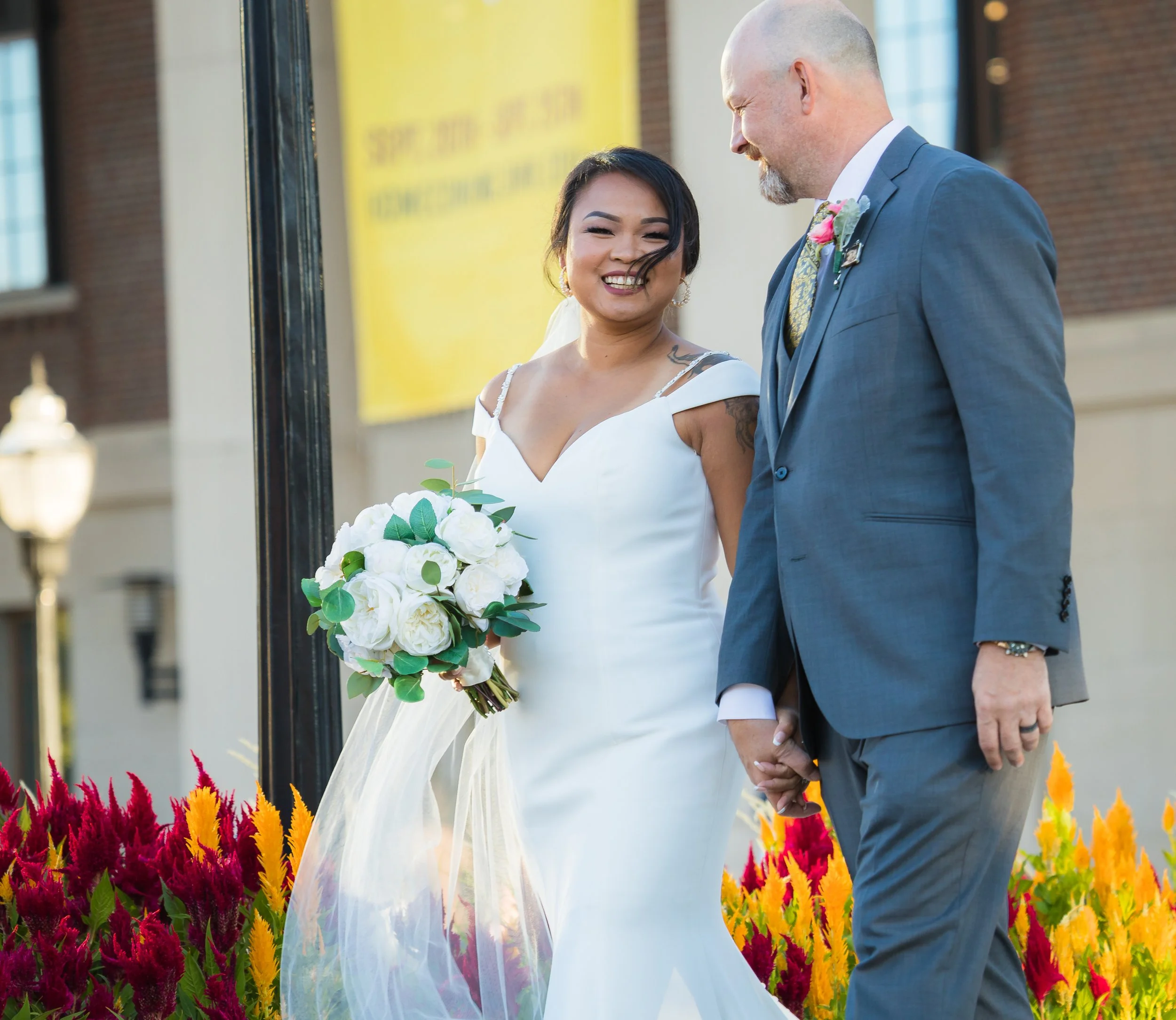 A bride and groom holding hands during their wedding ceremony outside. The bride is smiling and holding a bouquet of white flowers, while the groom is dressed in a blue suit with a boutonniere. Colorful flowers are in the foreground.