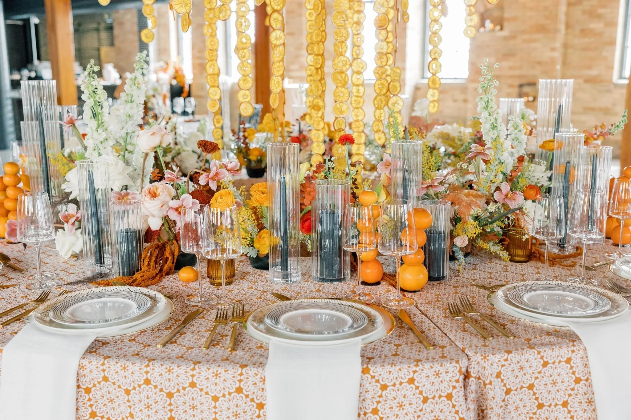 Decorated banquet table with floral arrangements, orange and yellow accents, glassware, and plates in an indoor setting for a celebration or event.
