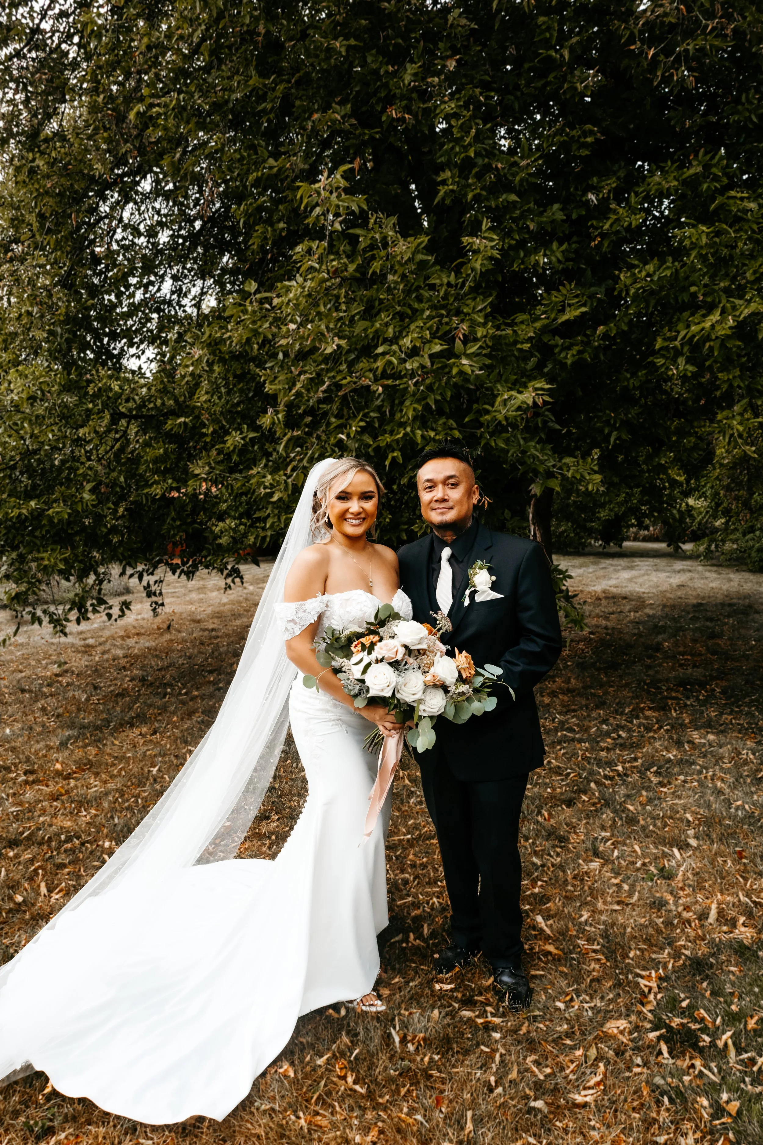 A bride in a white off-shoulder wedding gown holding a large bouquet and standing next to a groom in a black suit with a white shirt and black tie, both smiling, outdoors under a large tree with green leaves.