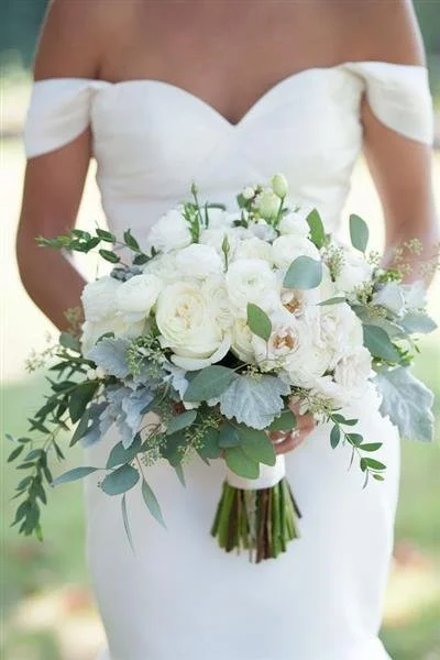 Bride in an off-shoulder white wedding dress holding a bouquet of white roses and greenery