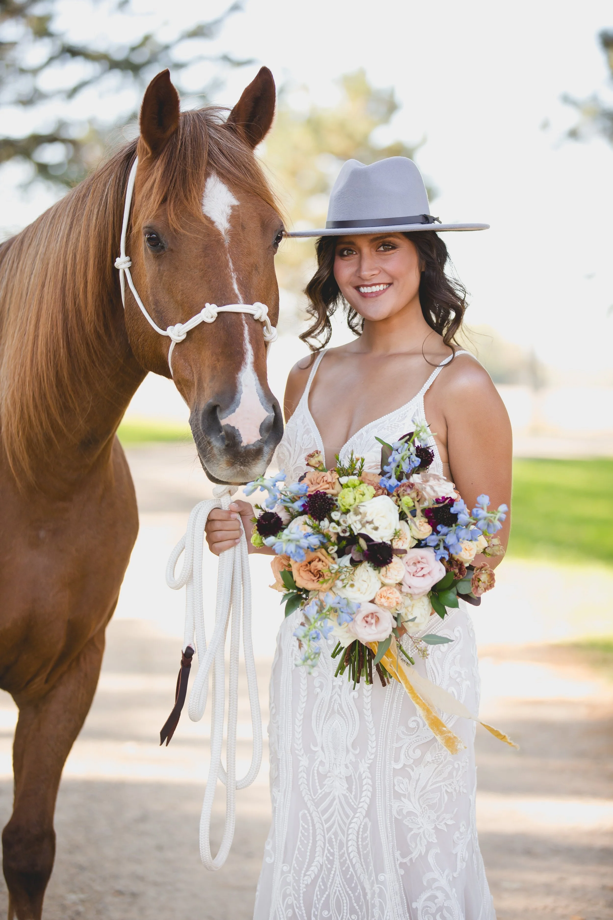 A woman in a white dress and a gray hat holding a bouquet of flowers standing next to a brown horse with a white stripe on its face outdoors.