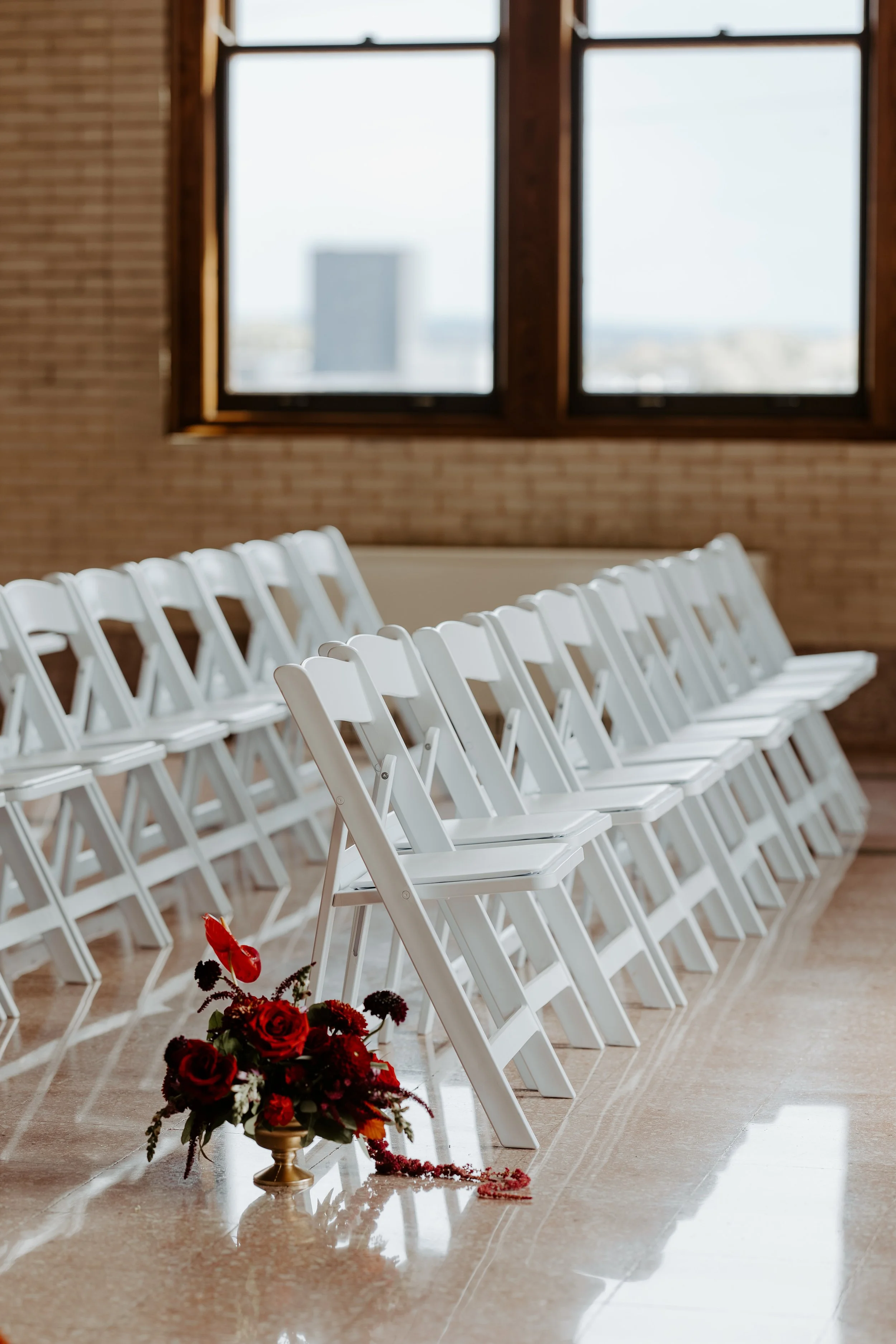 Rows of white folding chairs set up for a ceremony in a room with large windows, a brick wall, and a flower arrangement with red roses and greenery on the floor.