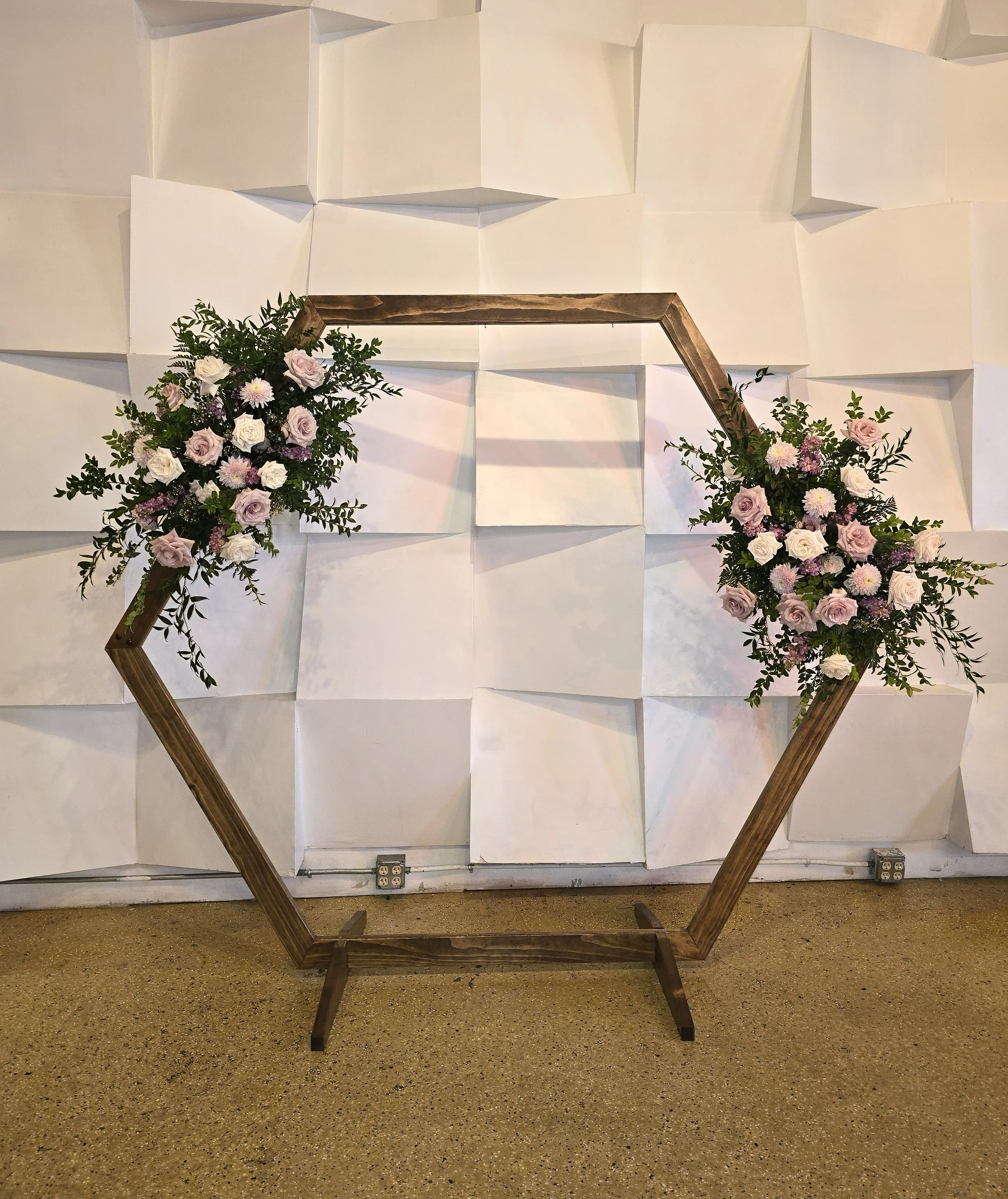 A floral arrangement of pink and white roses, daisies, and greenery displayed on a wooden geometric frame against a white, textured wall.