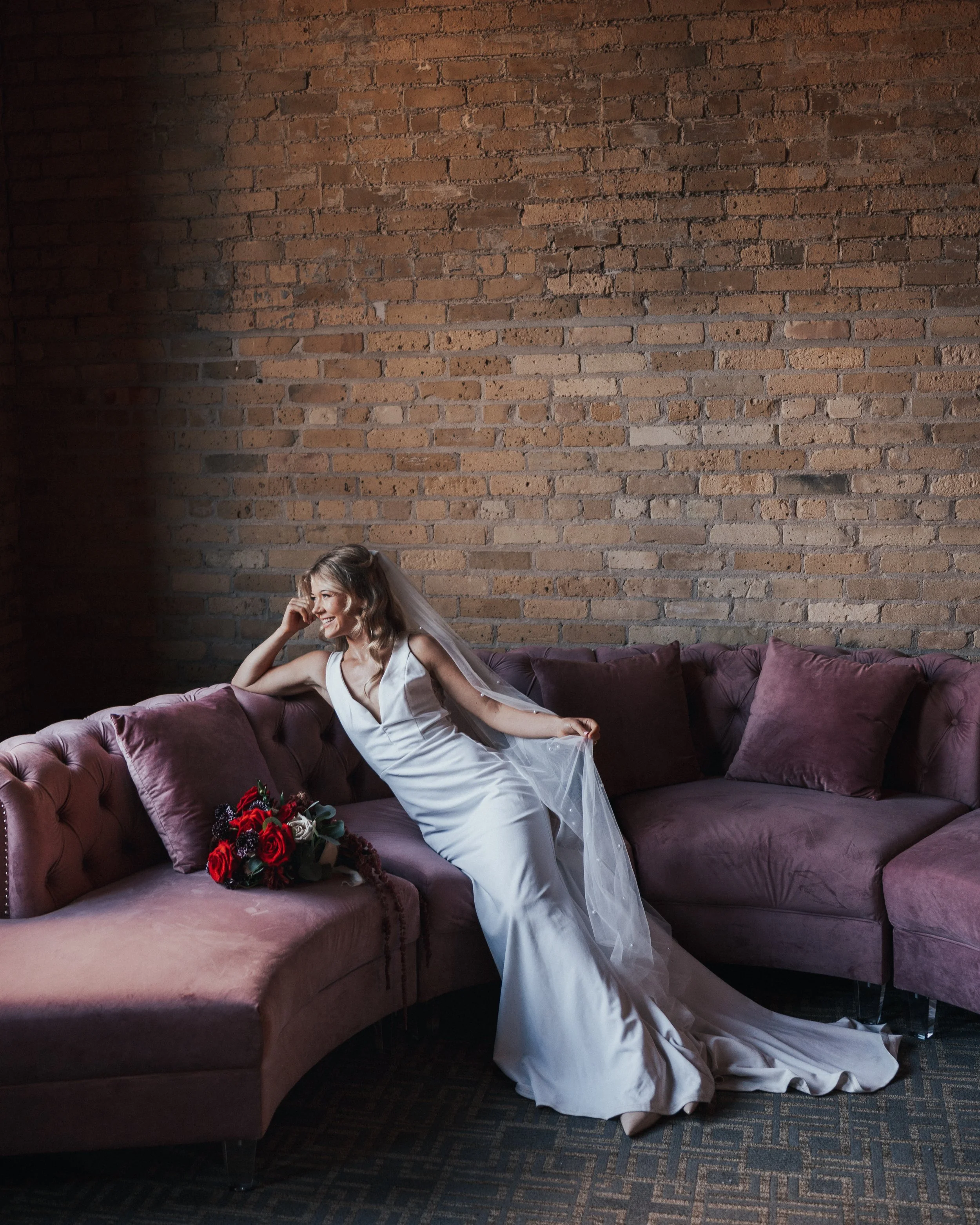 A woman in a wedding dress sitting on a purple velvet sofa with a bouquet of red and white roses beside her, smiling and looking to the side, in front of a brick wall.