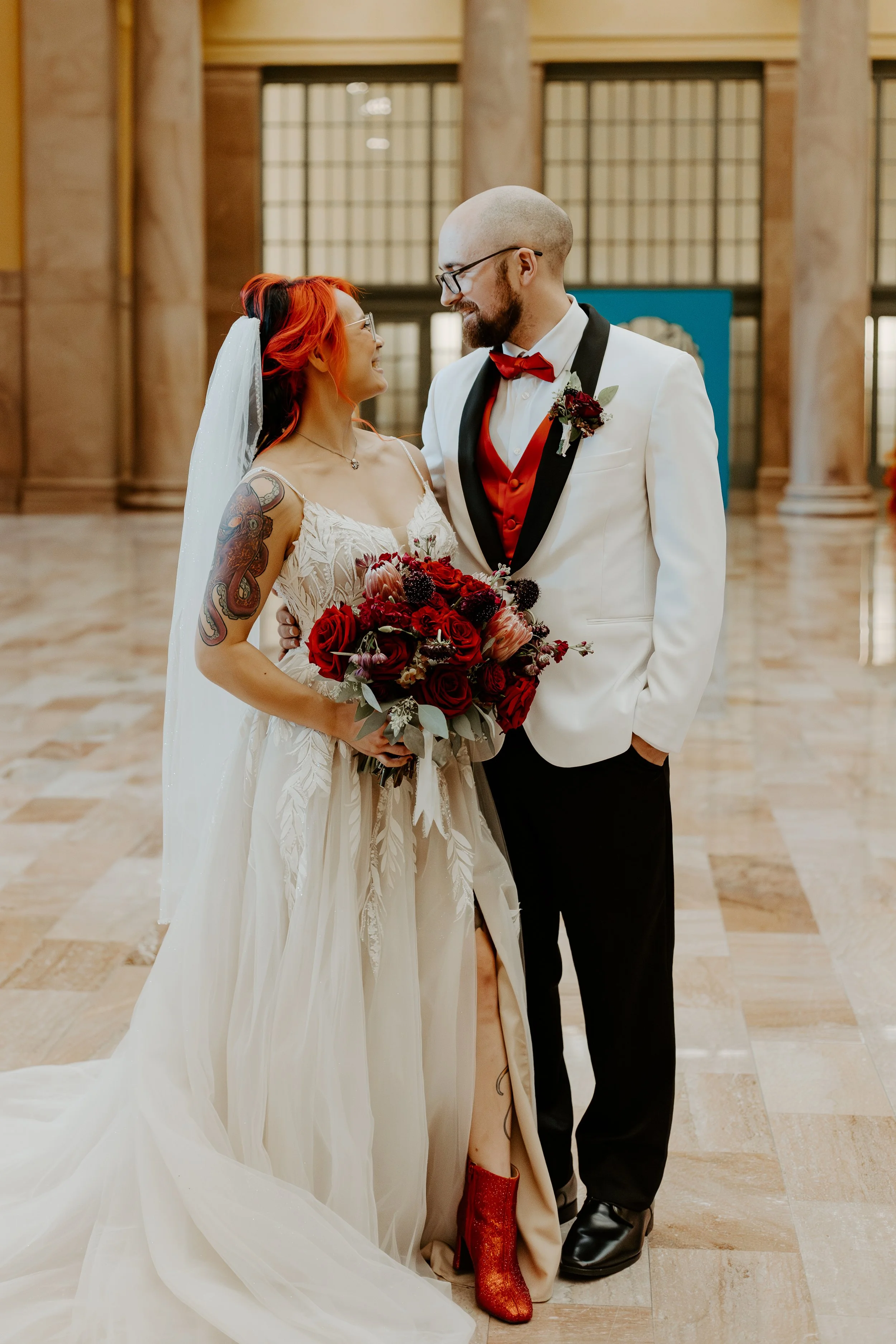A bride and groom looking at each other and smiling during their wedding in a grand hall with large windows and marble columns. The bride has red hair, tattoos on her arm, and is holding a bouquet of red and dark flowers. The groom is bald with a beard, wearing glasses and a white tuxedo with a red vest and bow tie.