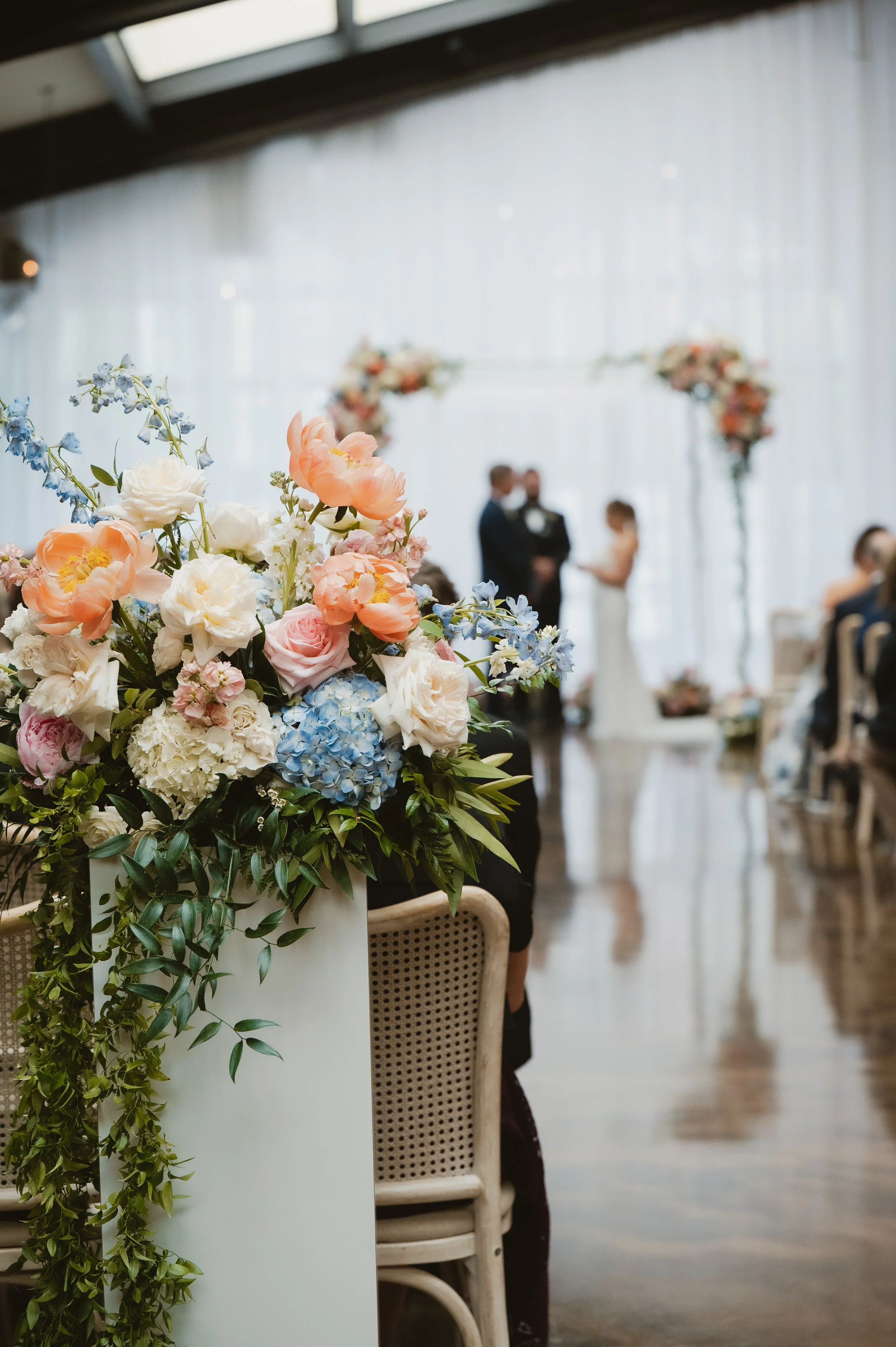 A wedding ceremony with a bouquet of pastel-colored flowers in the foreground and the bride and groom standing together in the background under floral arches, with guests seated nearby.