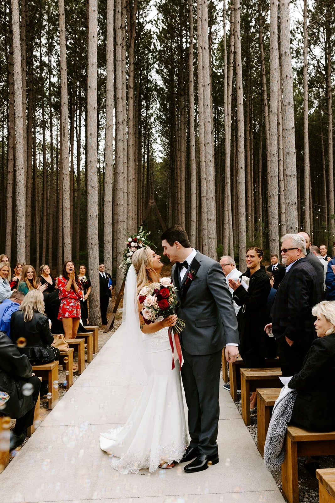 A bride and groom standing close together during a wedding ceremony in a wooded outdoor setting, surrounded by friends and family.