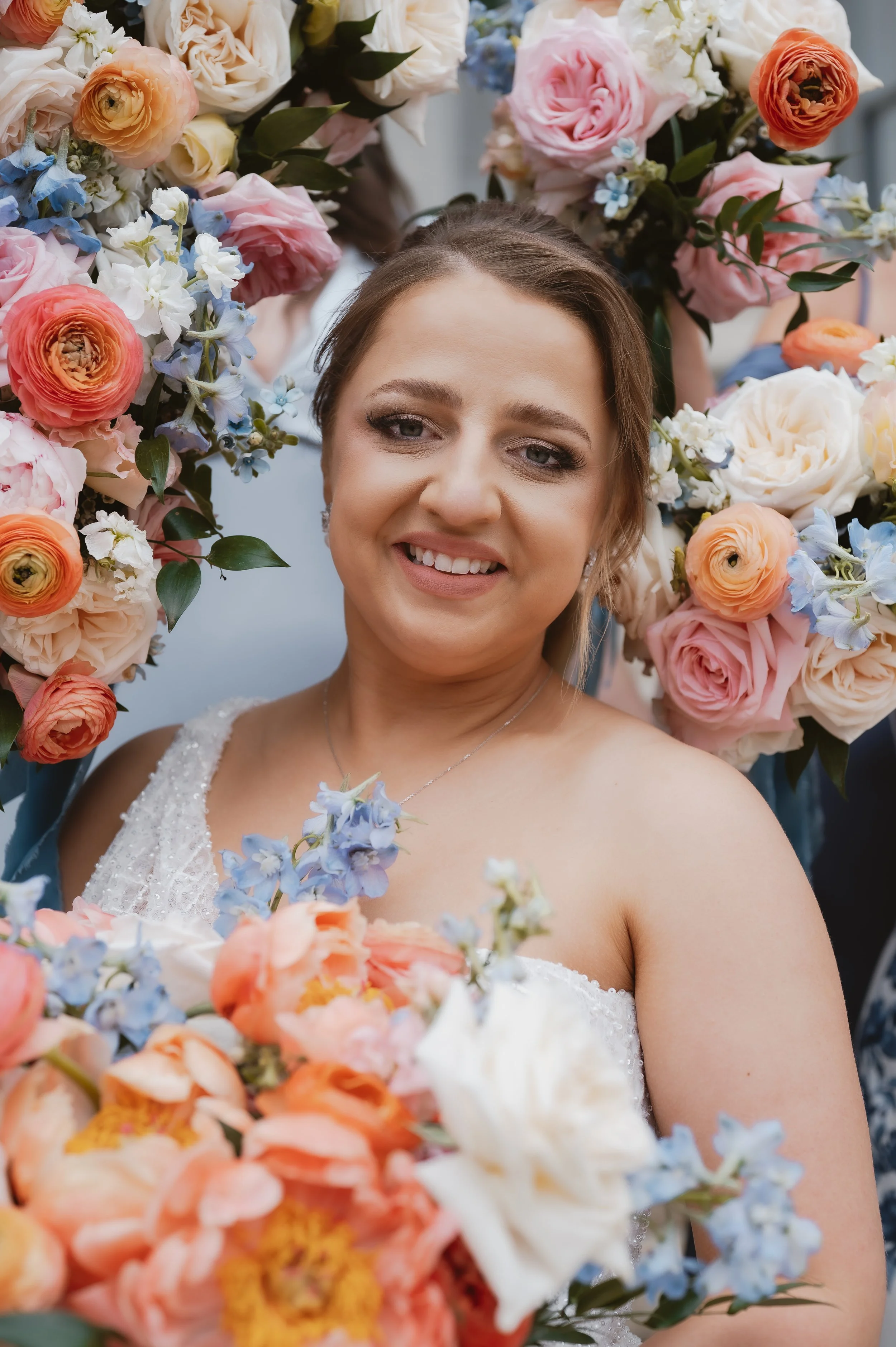 A smiling woman wearing a wedding dress surrounded by an arch of colorful flowers including roses, peonies, and other blossoms.