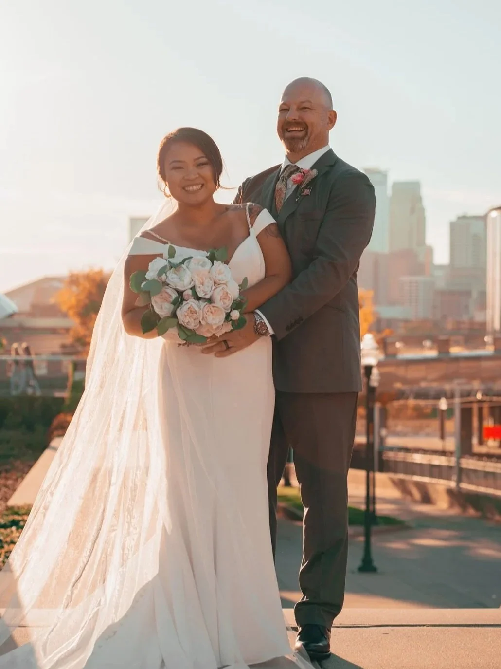 A happy bride and groom standing outdoors in a cityscape at sunset, with the bride holding a bouquet of blush pink roses and wearing a white wedding gown with a veil, and the groom in a dark suit.