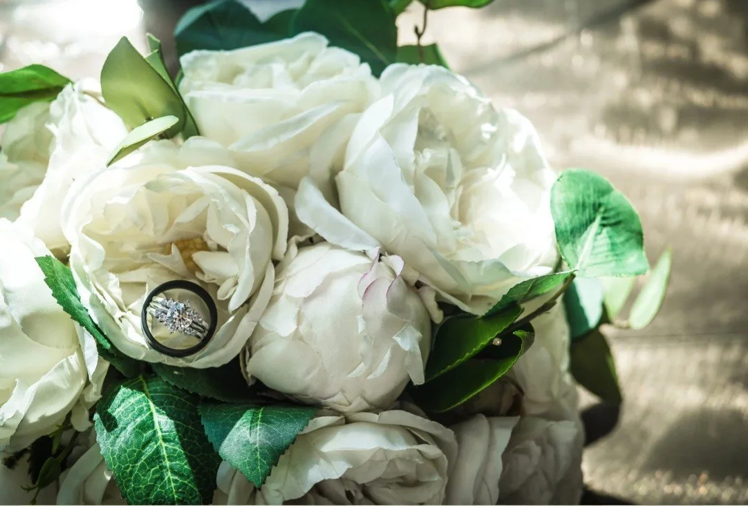 Close-up of white roses with a diamond engagement ring resting on one of the blooms, surrounded by green leaves.