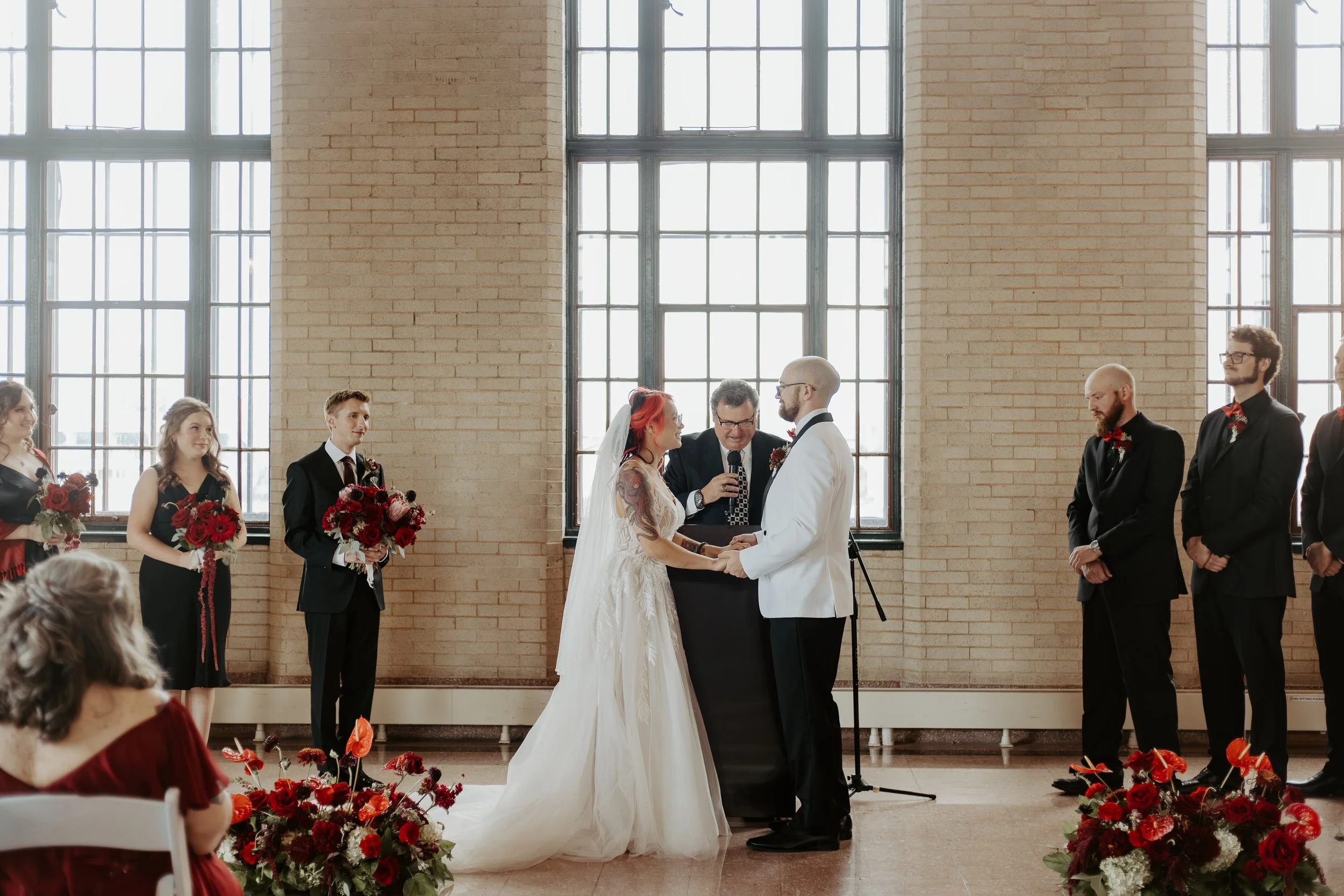 A wedding ceremony with a bride and groom exchanging vows in front of an officiant, surrounded by bridesmaids and groomsmen in formal attire, inside a space with large windows and brick walls, decorated with red flowers.