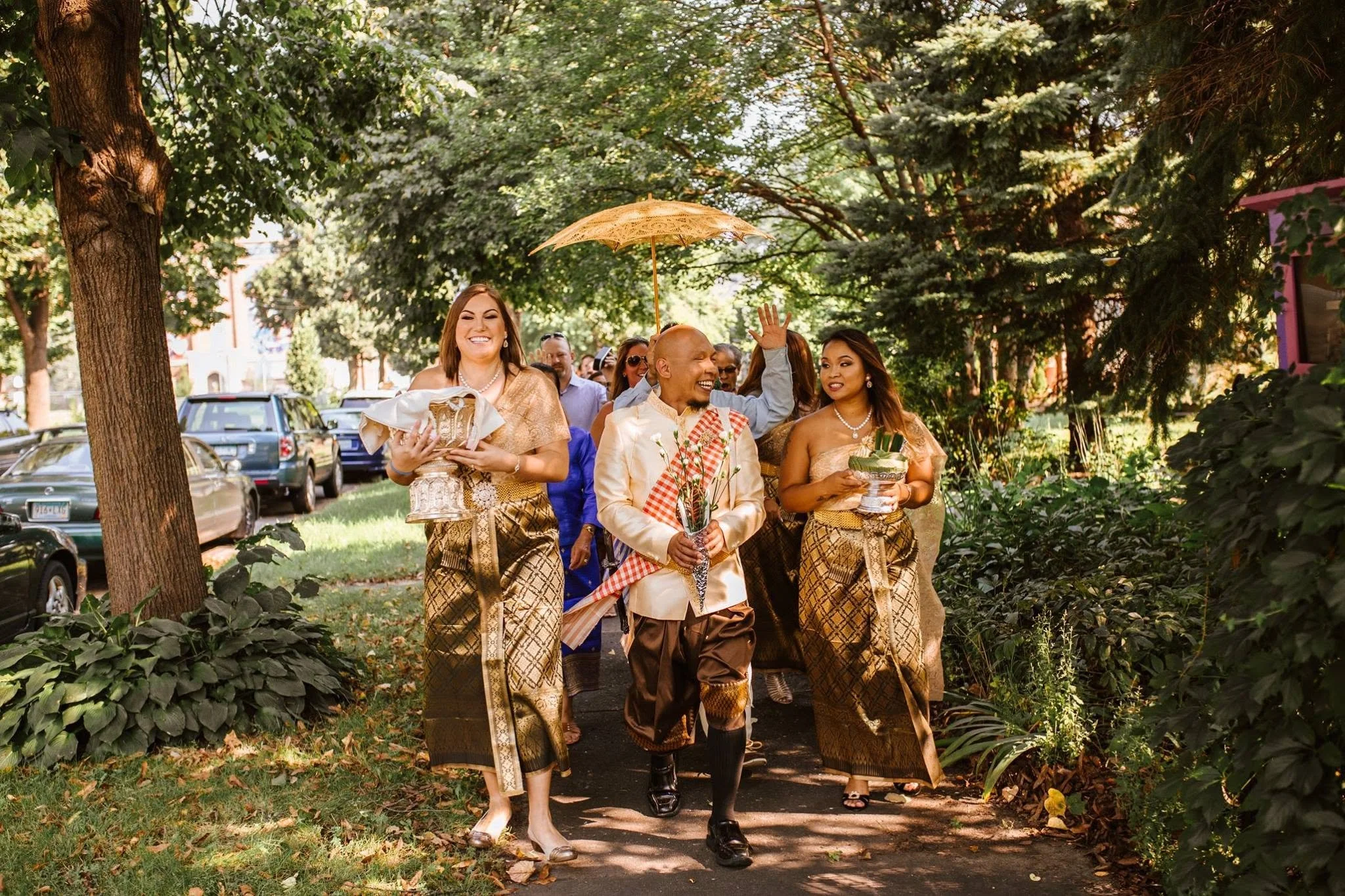 A group of people dressed in traditional attire walking through a park with trees, holding gifts and an umbrella, smiling and appearing happy.